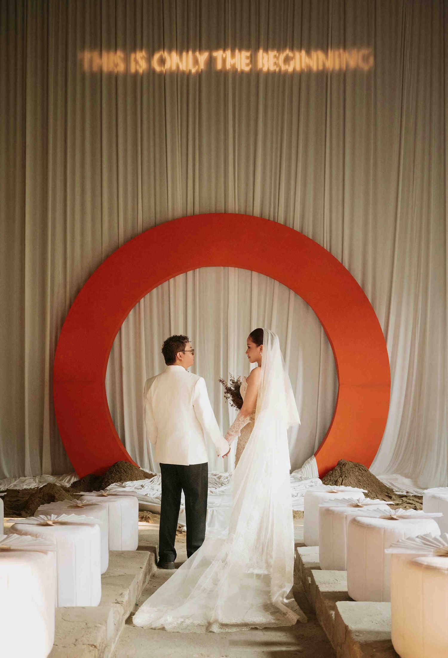 Couple stands before a red circular arch at a wedding ceremony with neon sign reading THIS IS ONLY THE BEGINNING.