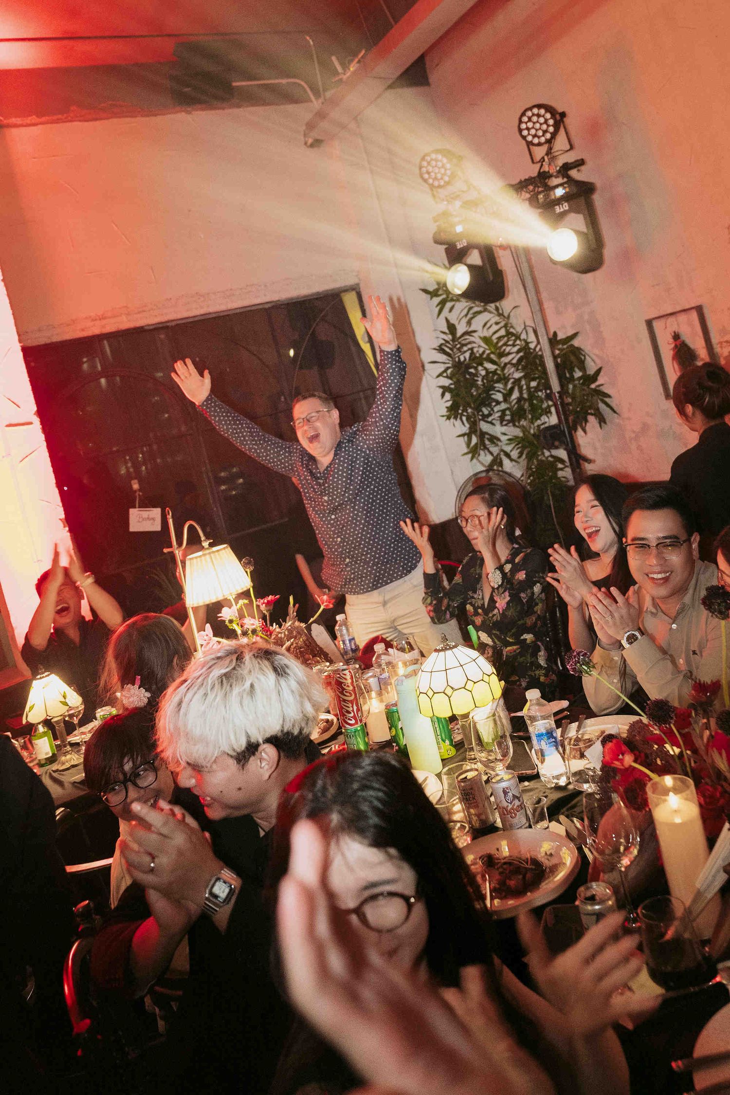 Excited guests celebrate at a lively party with dramatic stage lighting and a long table filled with drinks.