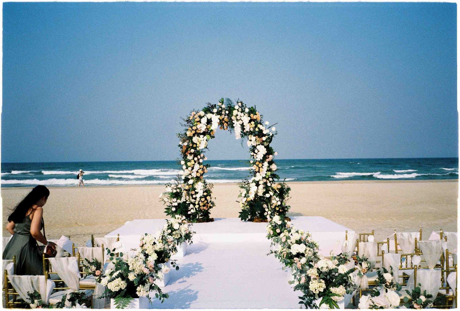 Elegant beach wedding ceremony setup with floral arch, white aisle runner, and flower arrangements by the ocean.