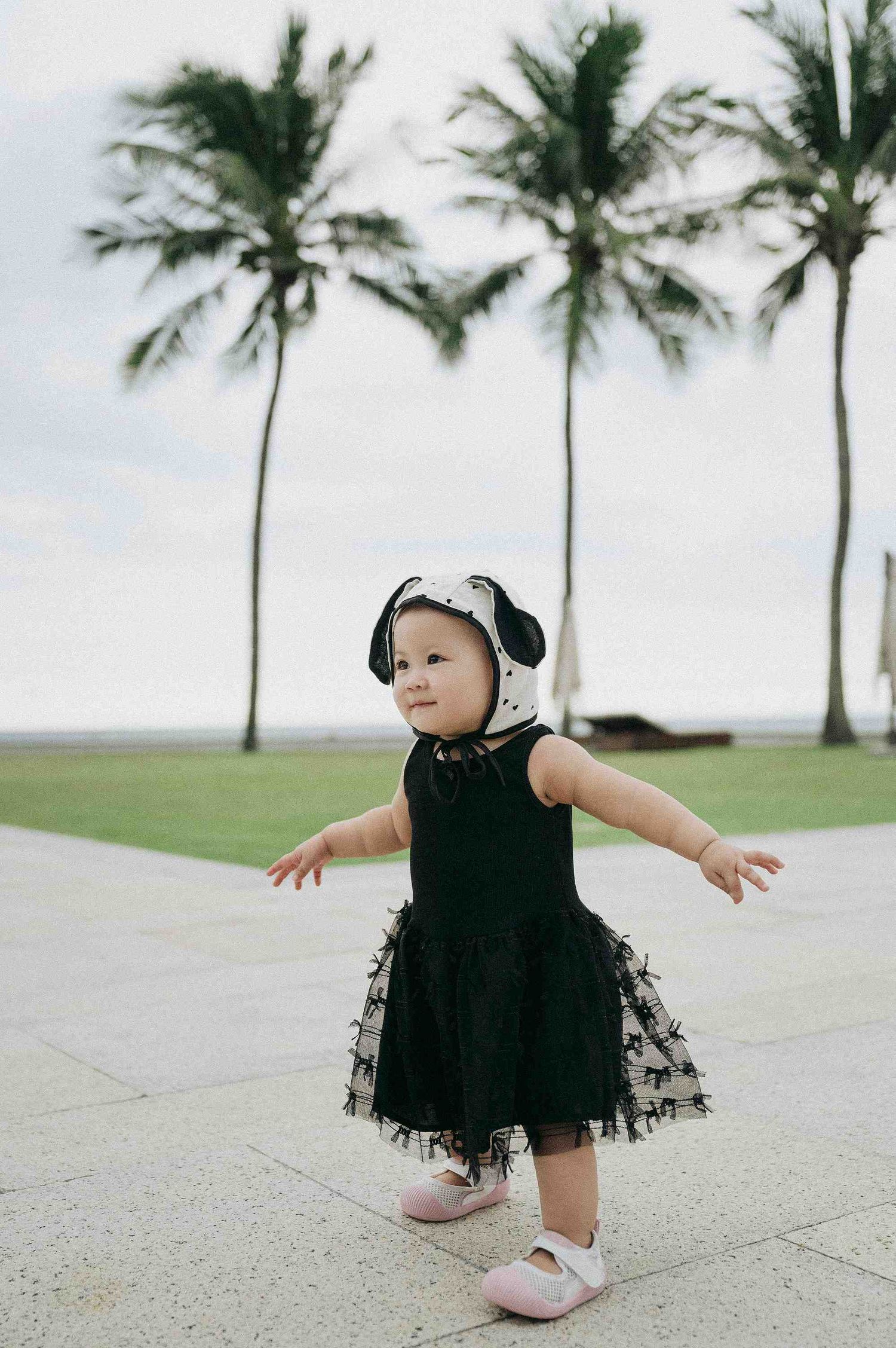 Da Nang family: small child in a black dress plays on a concrete path near palm trees, by AnhVuWedding.