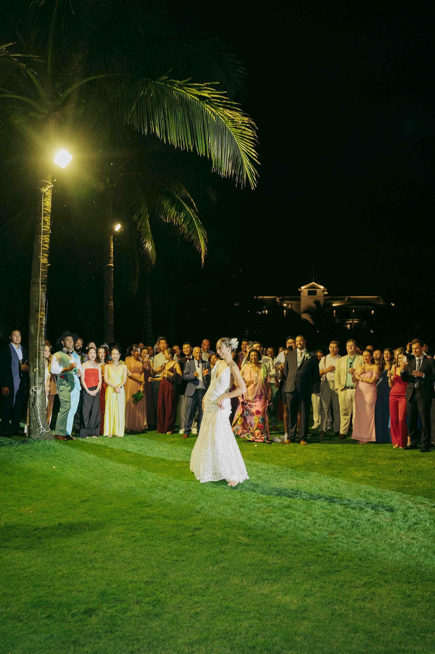 Bride in white gown prepares to toss bouquet to guests at outdoor tropical wedding reception at night.