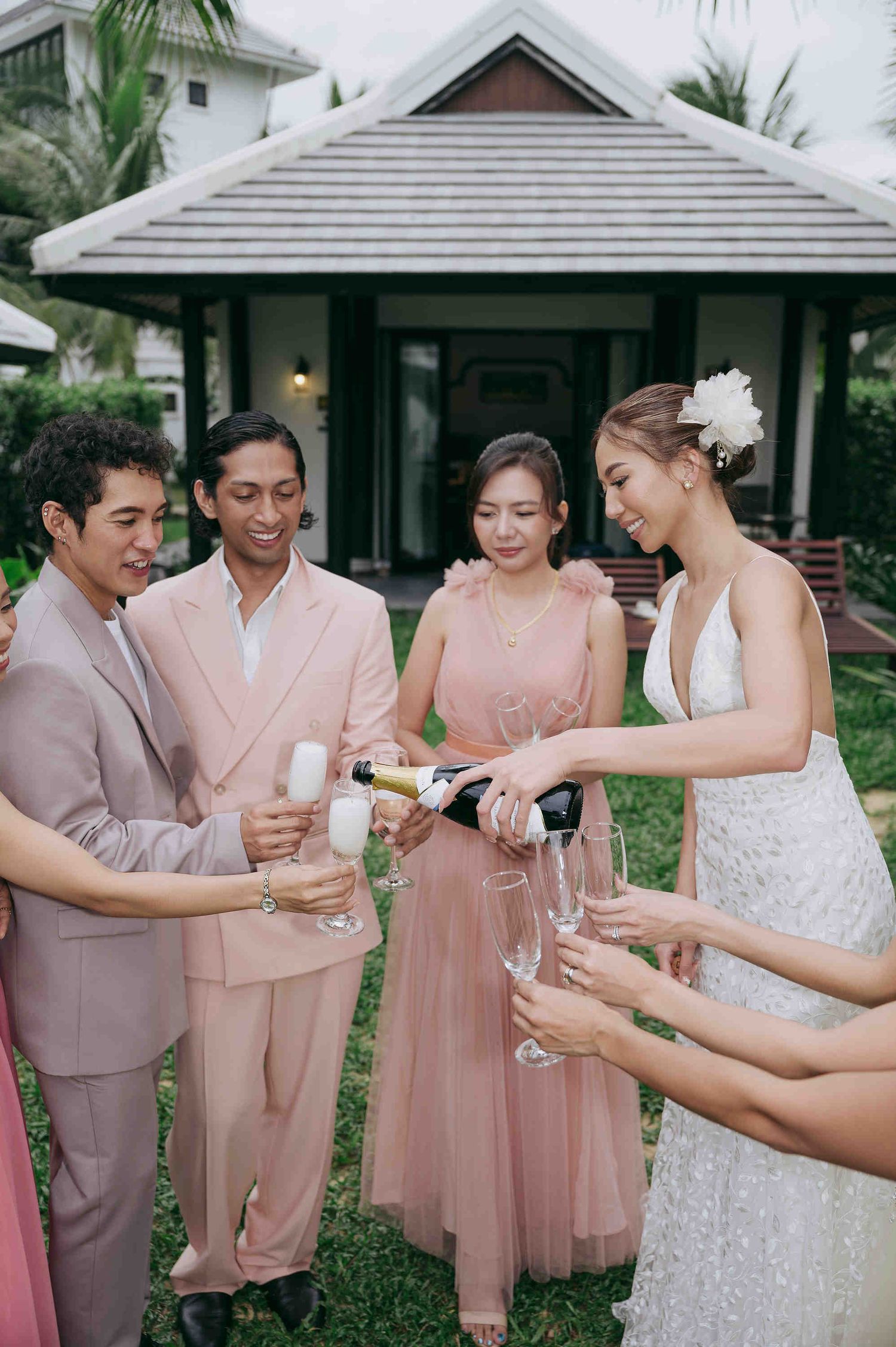 Bride pouring champagne for guests in pink attire during outdoor wedding celebration near tropical villa.