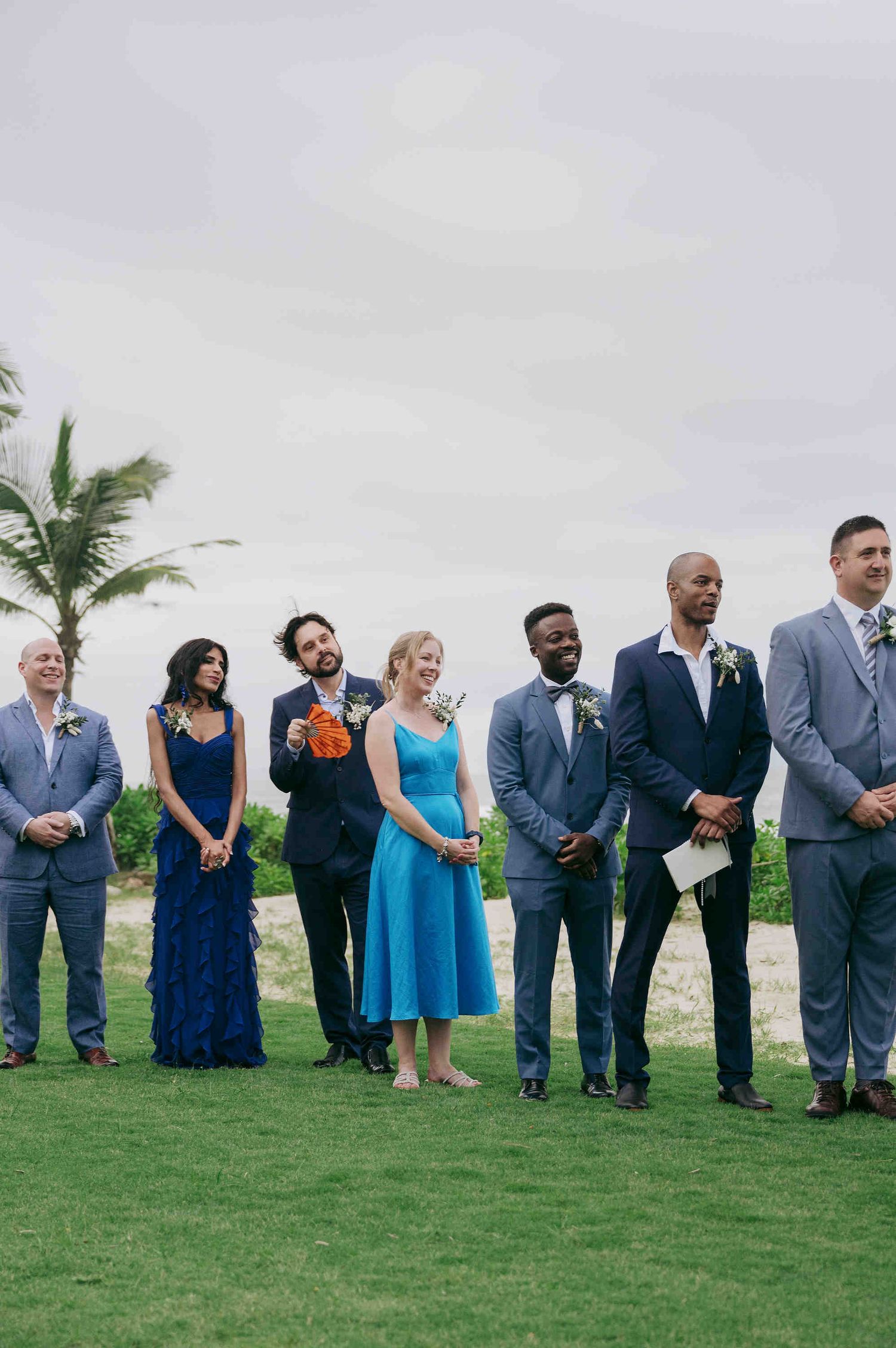 Wedding party in blue attire standing on green lawn with palm trees, featuring bridesmaids and groomsmen at outdoor ceremony.