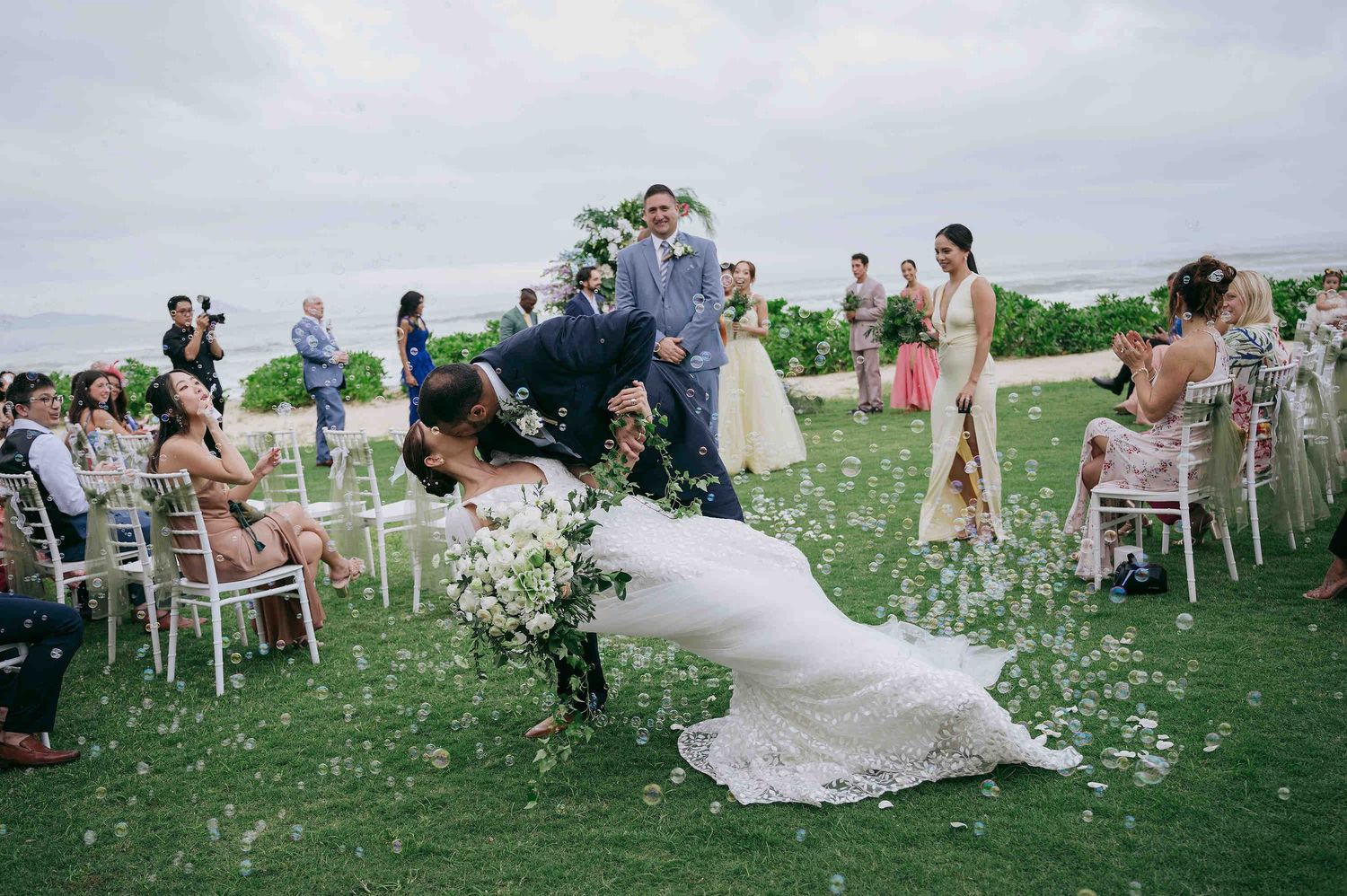 Groom dips bride during outdoor wedding ceremony on lush green lawn with white chairs and floral petals.