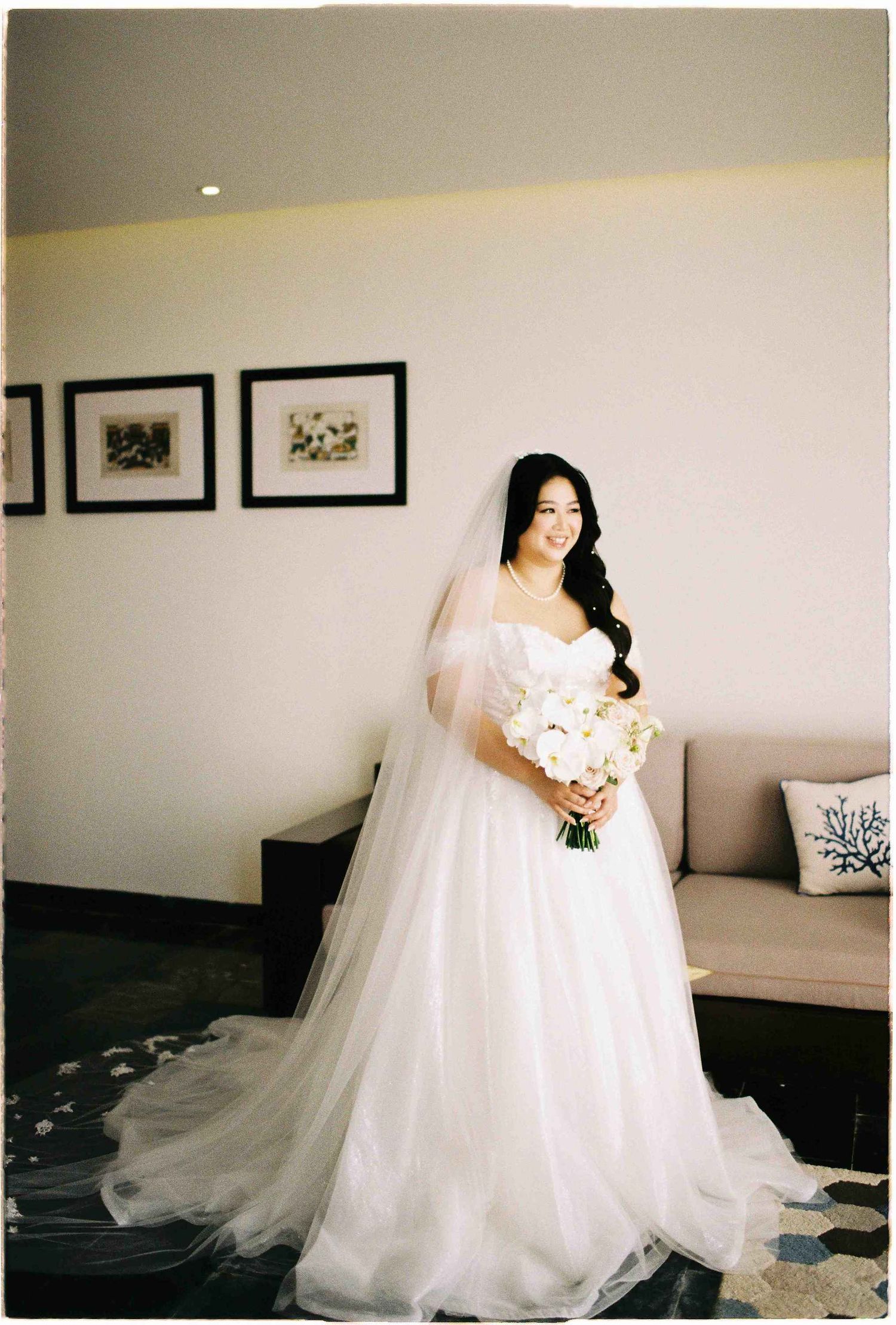 Bride in a white ballgown with veil holding a white bouquet, standing in an elegant room before her wedding.