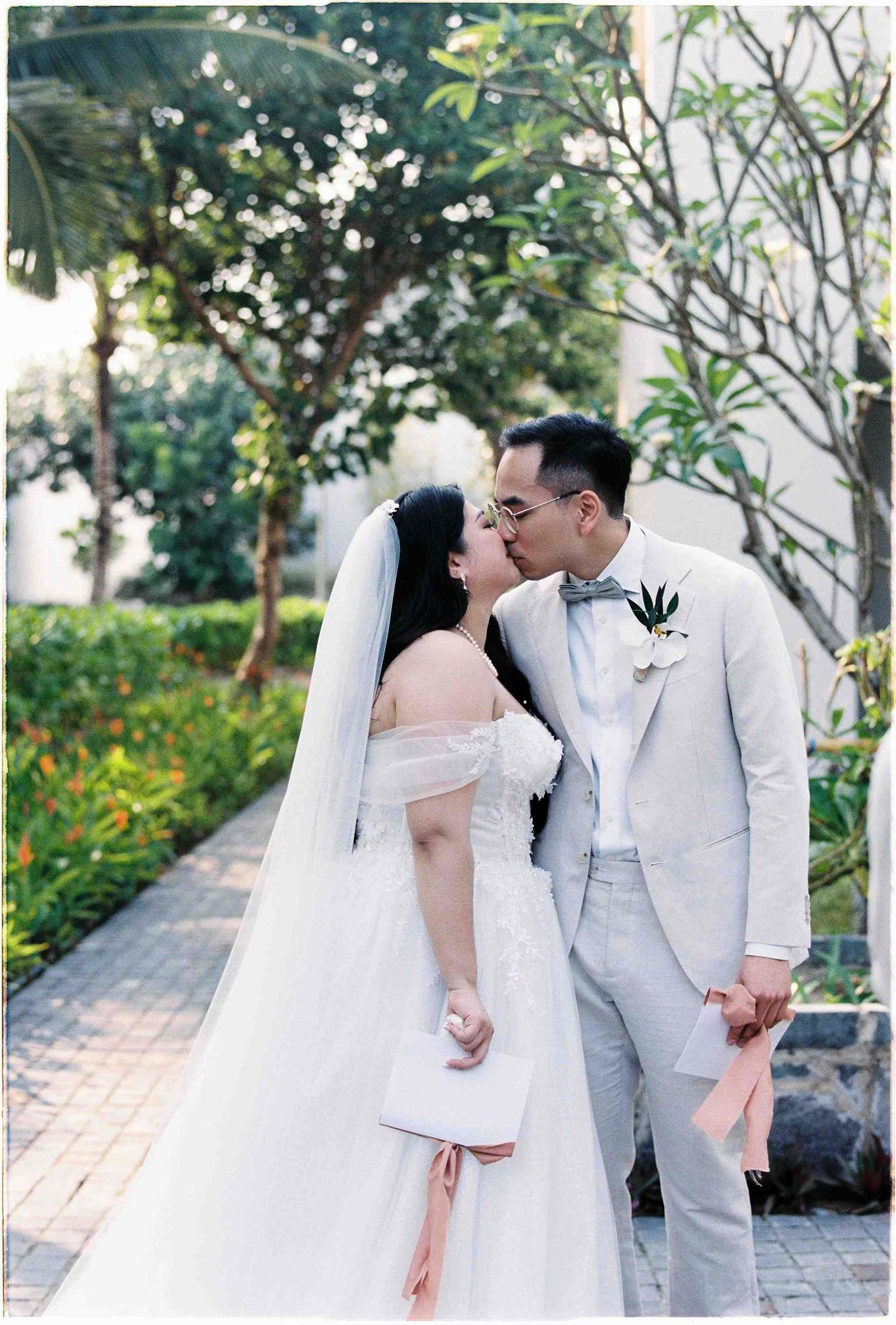 Bride and groom share a romantic kiss outdoors, she in a white off-shoulder gown with veil, he in a light gray suit.