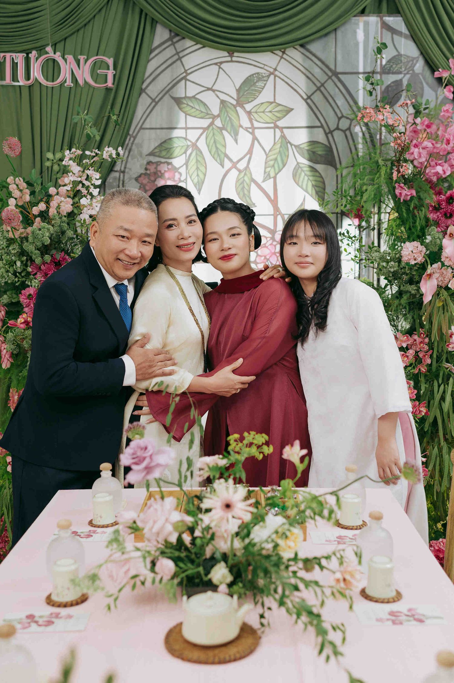 Family of four posing together at a floral Vietnamese wedding celebration with pink and green decorations.