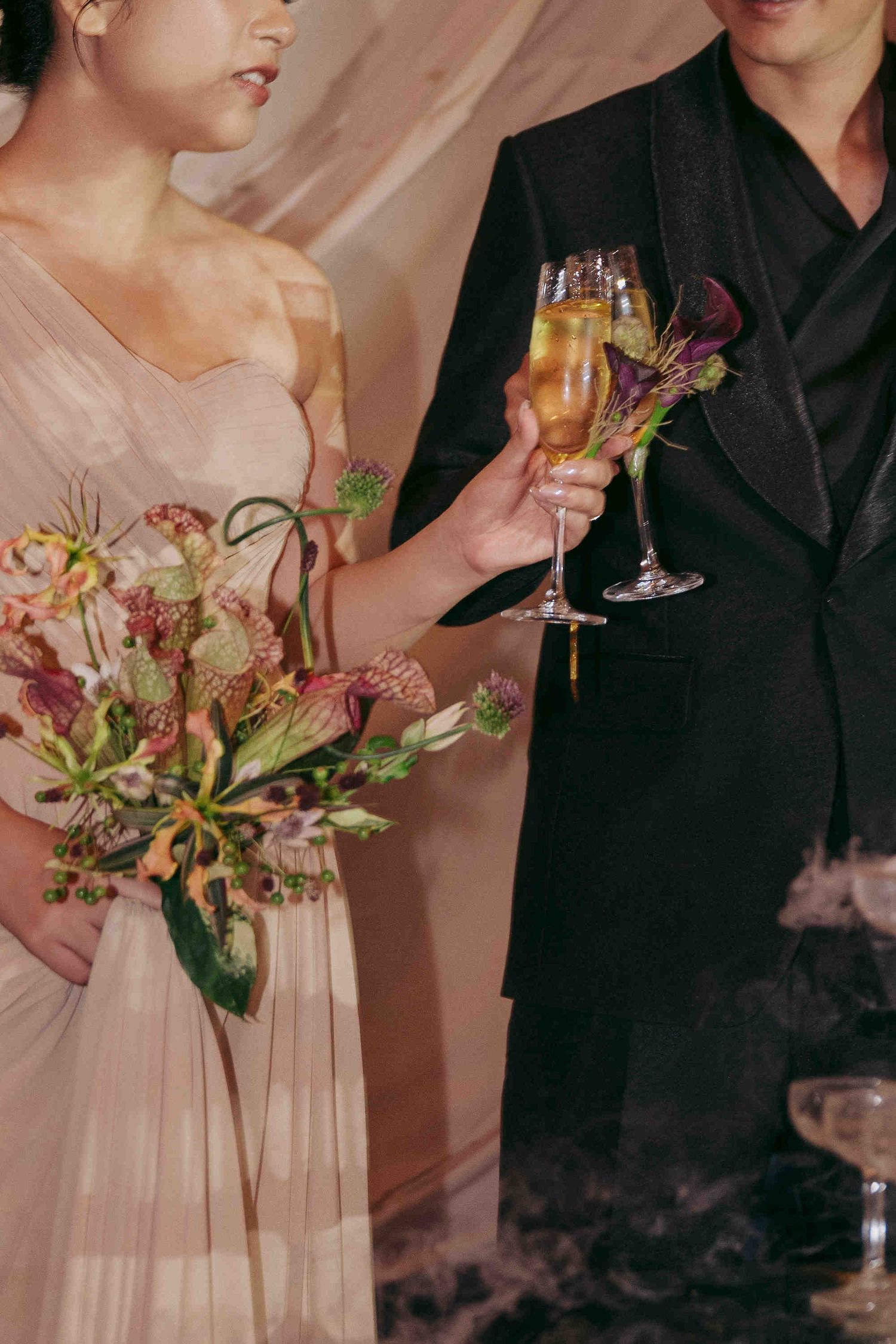 Bride in blush gown and groom in black suit clinking champagne flutes at wedding reception with floral bouquet.
