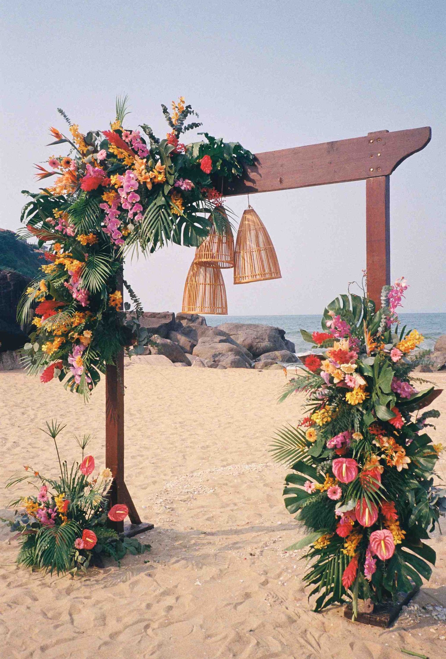Lang Co wedding: wooden arch decorated with vibrant tropical flowers and hanging lanterns on a beach, by AnhVuWedding.