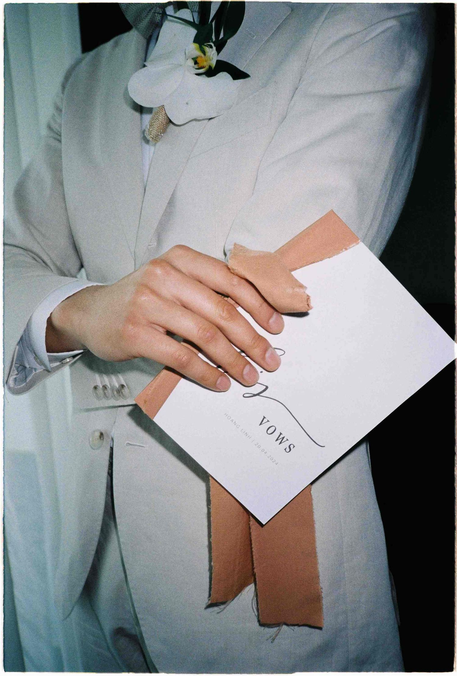 Groom in white suit holding wedding vows booklet with peach ribbon boutonniere at ceremony.
