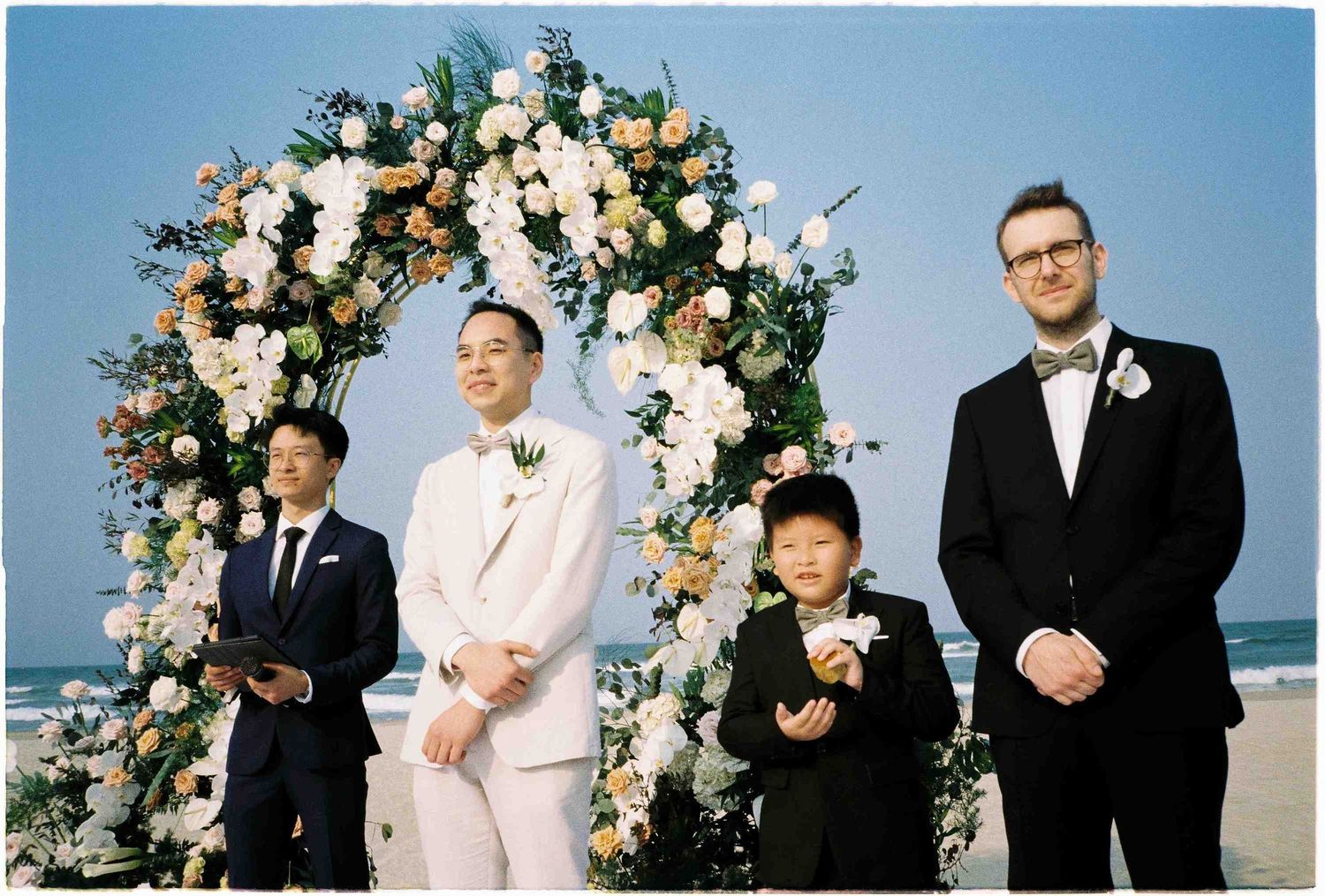 Two grooms and a young ring bearer pose under a floral arch at a beachside wedding ceremony.