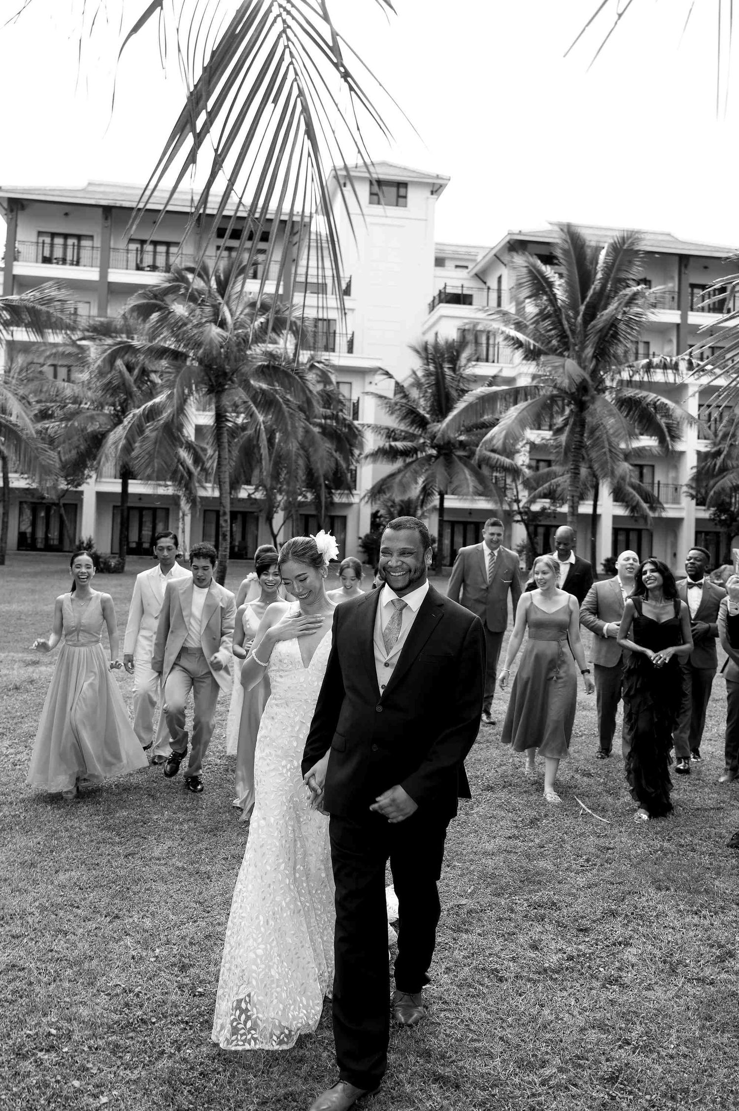 Black and white photo of a smiling bride and groom leading their wedding party across a tropical lawn with palm trees.