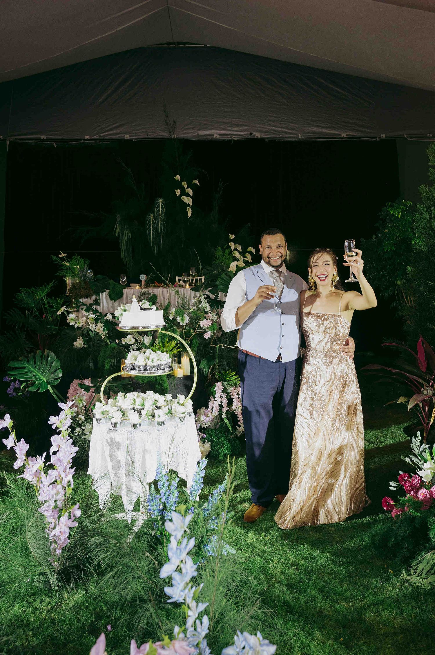 Bride in gold gown and groom toast beside wedding cake surrounded by lush tropical florals at outdoor night reception.