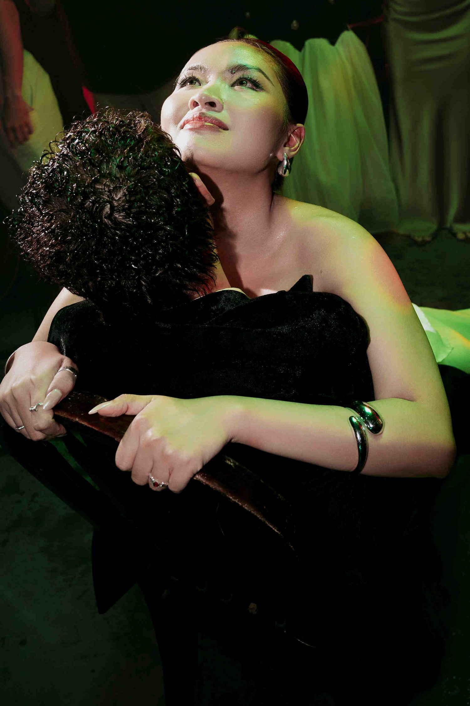 A woman in a black strapless dress embraces a person with curly hair under dramatic green stage lighting.