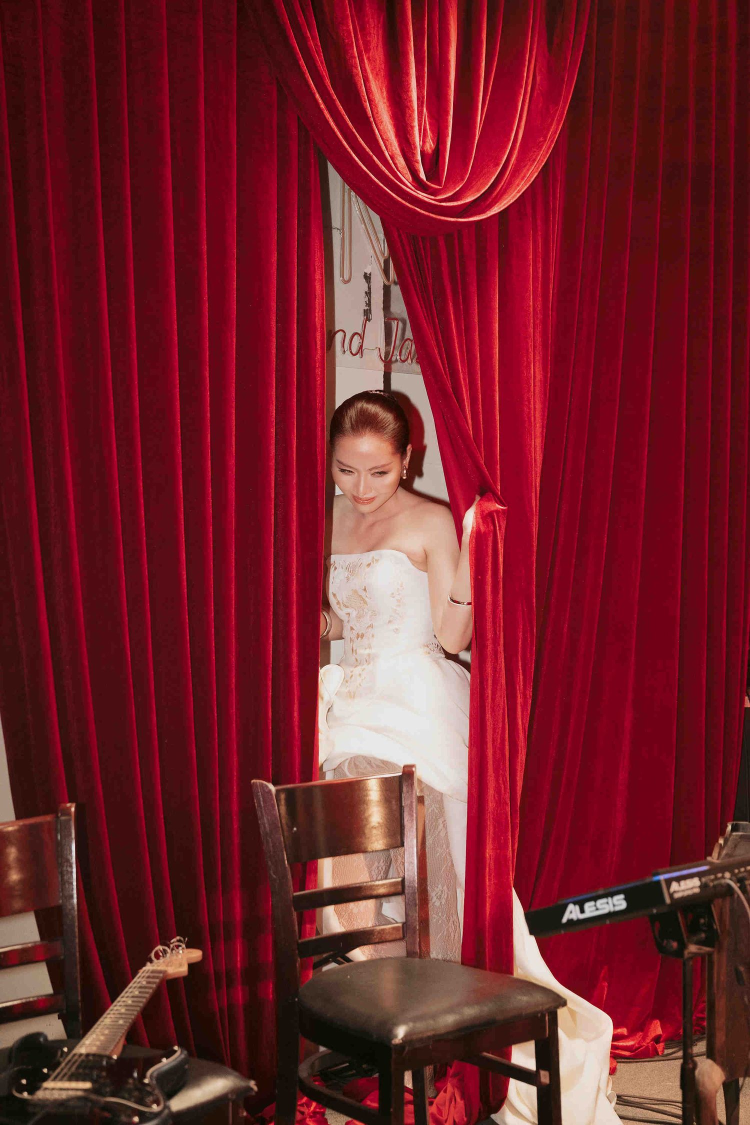 Bride in white strapless gown emerging through red velvet curtains near musical instruments on stage.