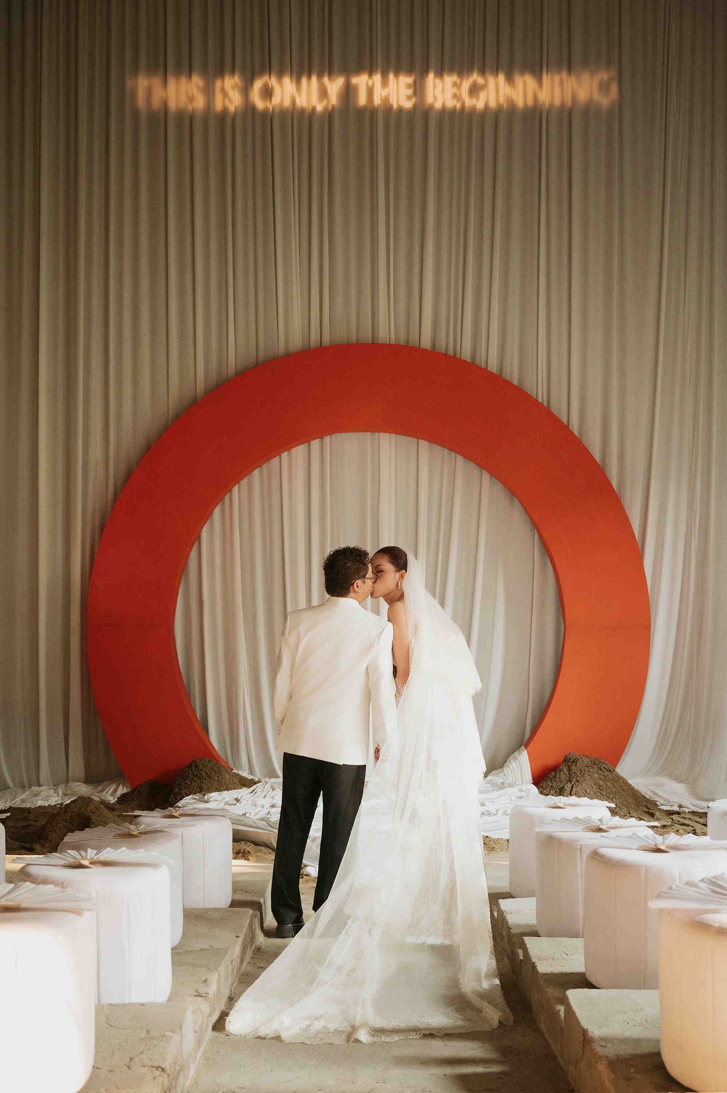 Bride and groom kissing under red circular arch with neon sign reading This Is Only The Beginning at wedding reception.
