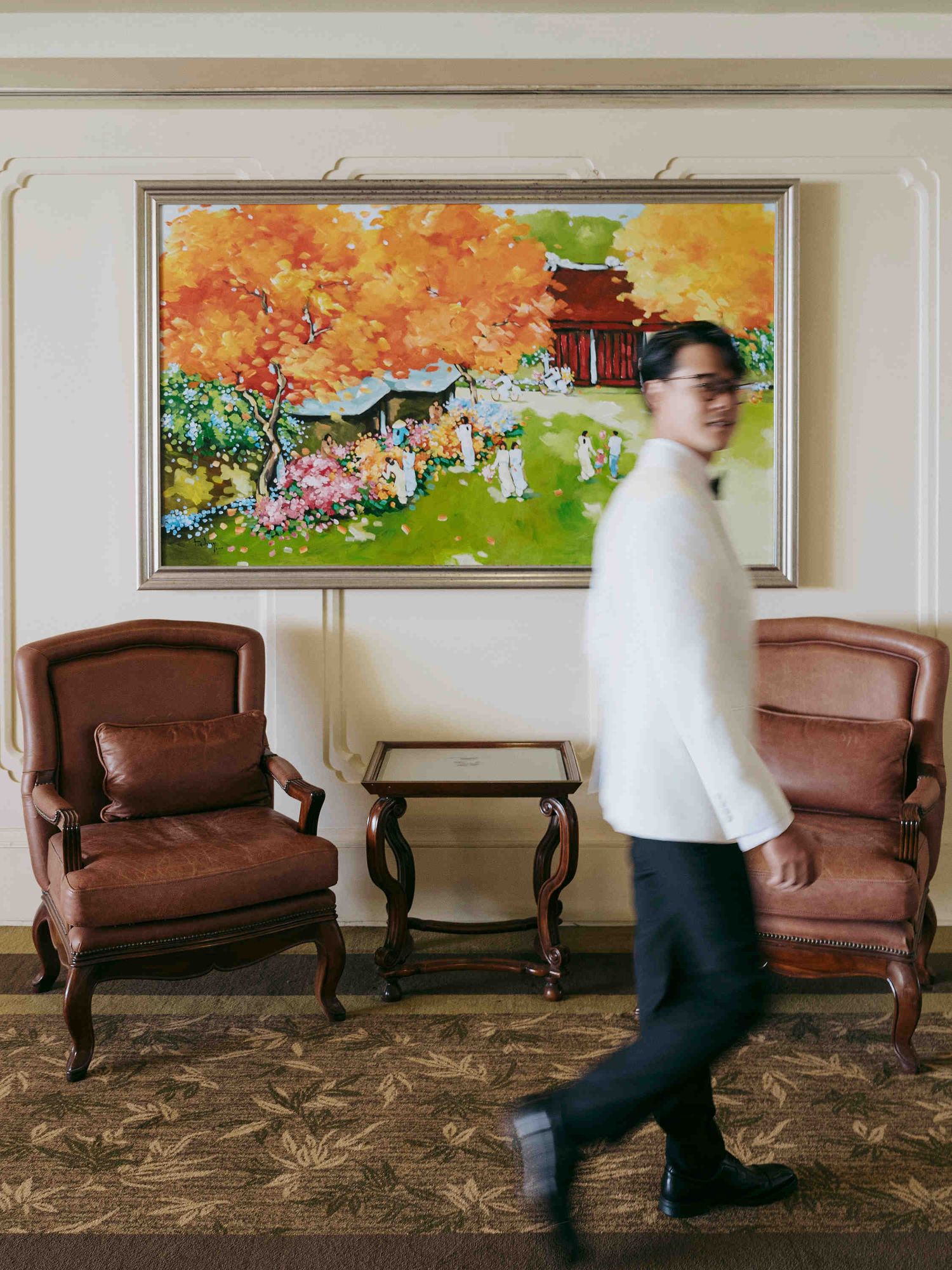 Man walking past two leather armchairs and a vibrant autumn landscape painting in an elegant hotel lobby.