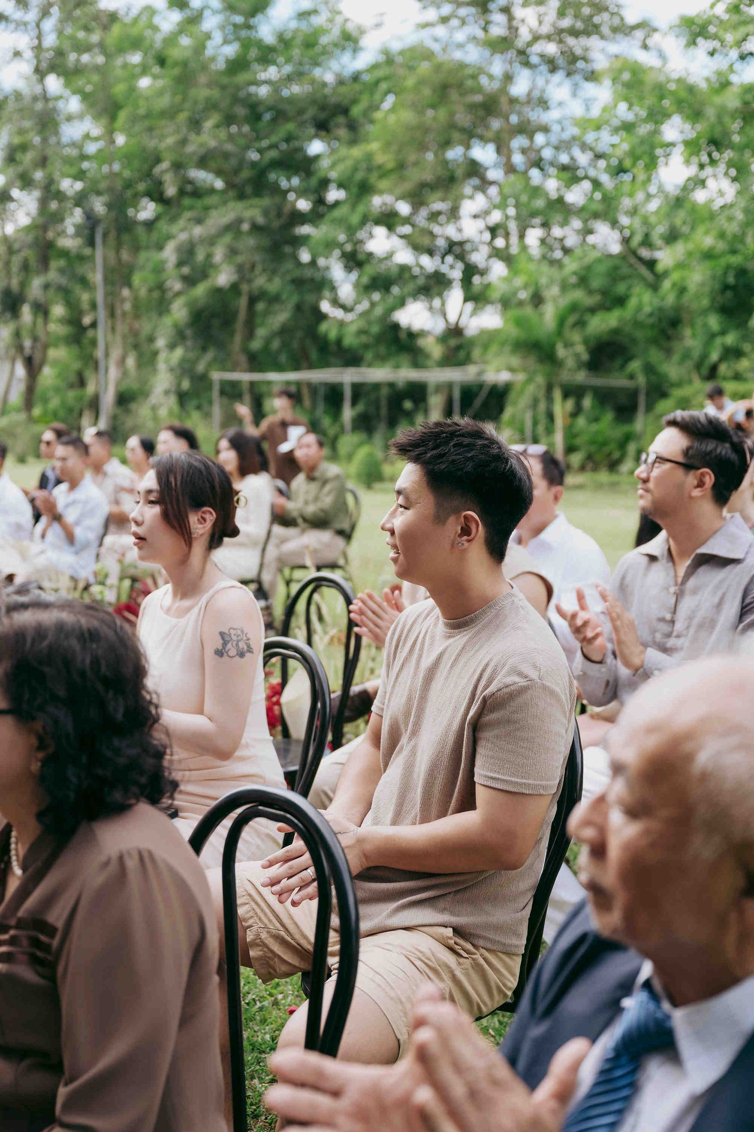 Guests clapping and smiling during an outdoor wedding ceremony seated on black chairs surrounded by lush greenery.