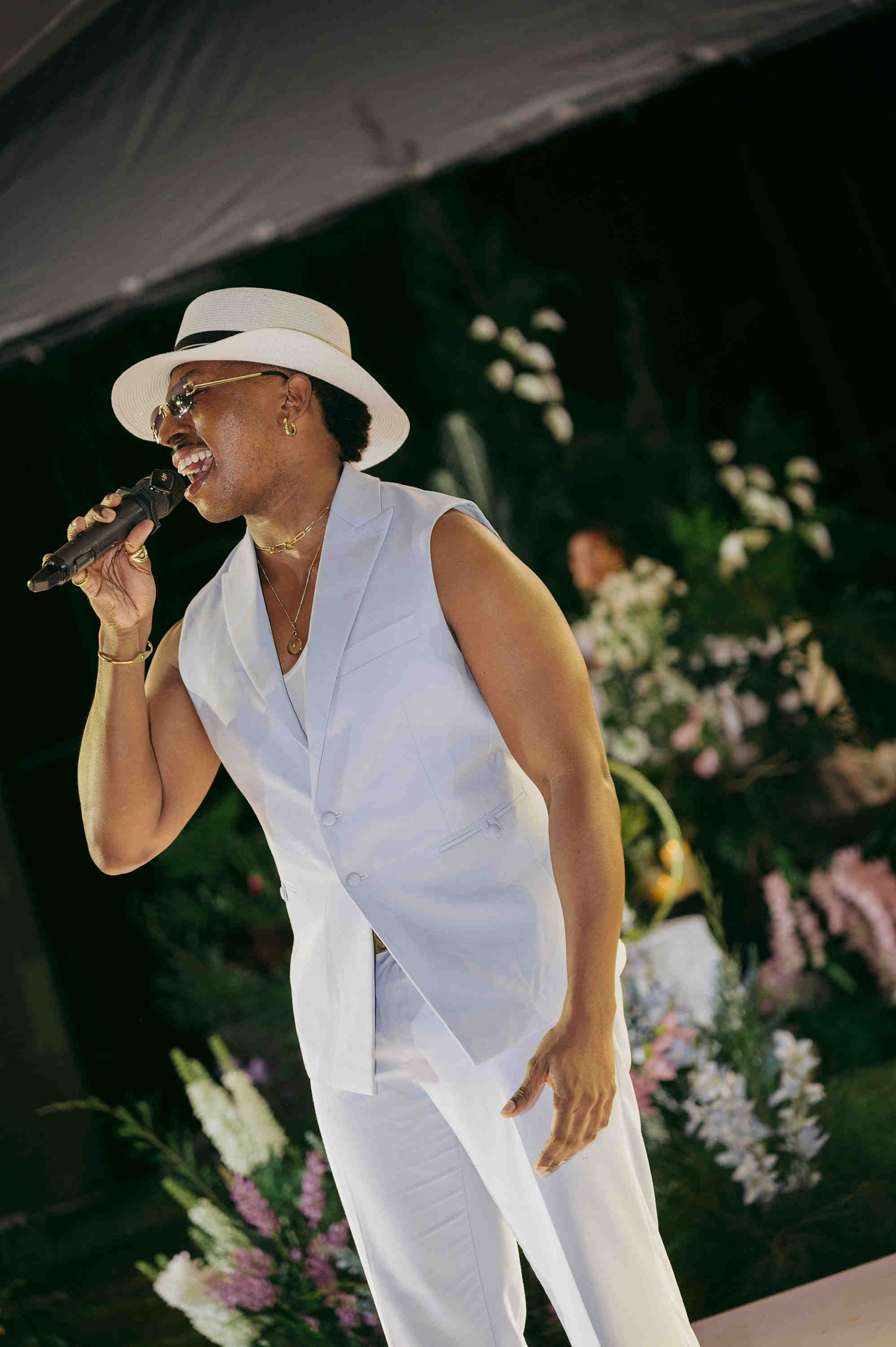 Elegant performer in white suit and hat singing into microphone at an outdoor floral wedding celebration.