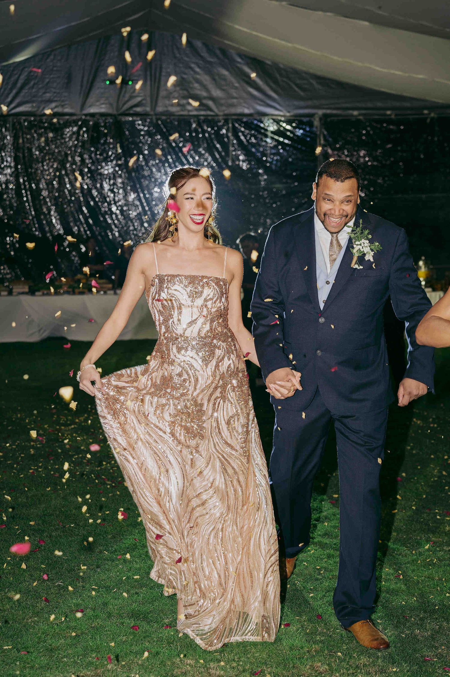Bride in gold sequin gown and groom in navy suit walk through confetti shower at elegant outdoor wedding reception.
