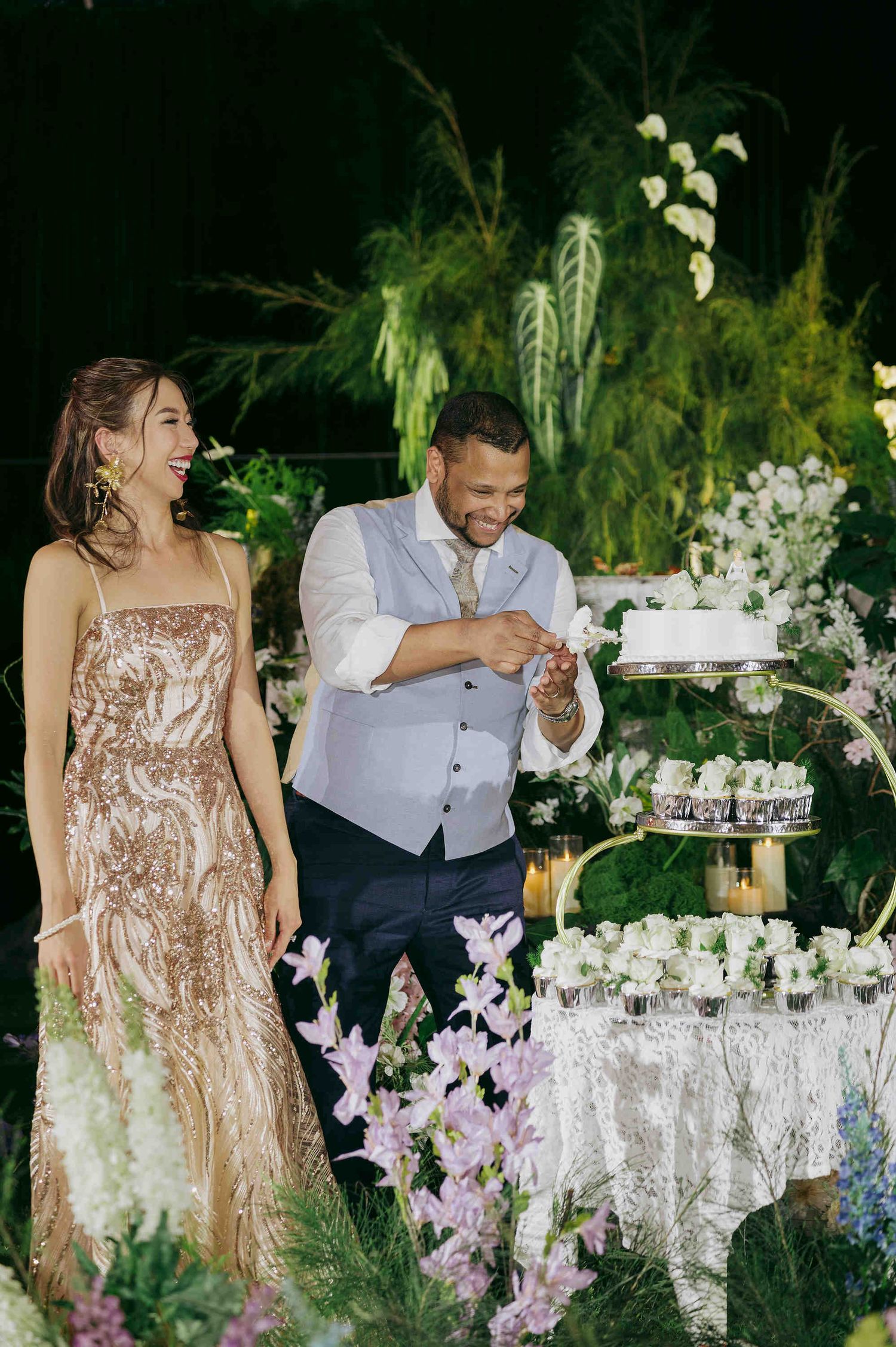 Couple laughing together beside elegant wedding cake display surrounded by lush greenery and purple flowers at outdoor reception.