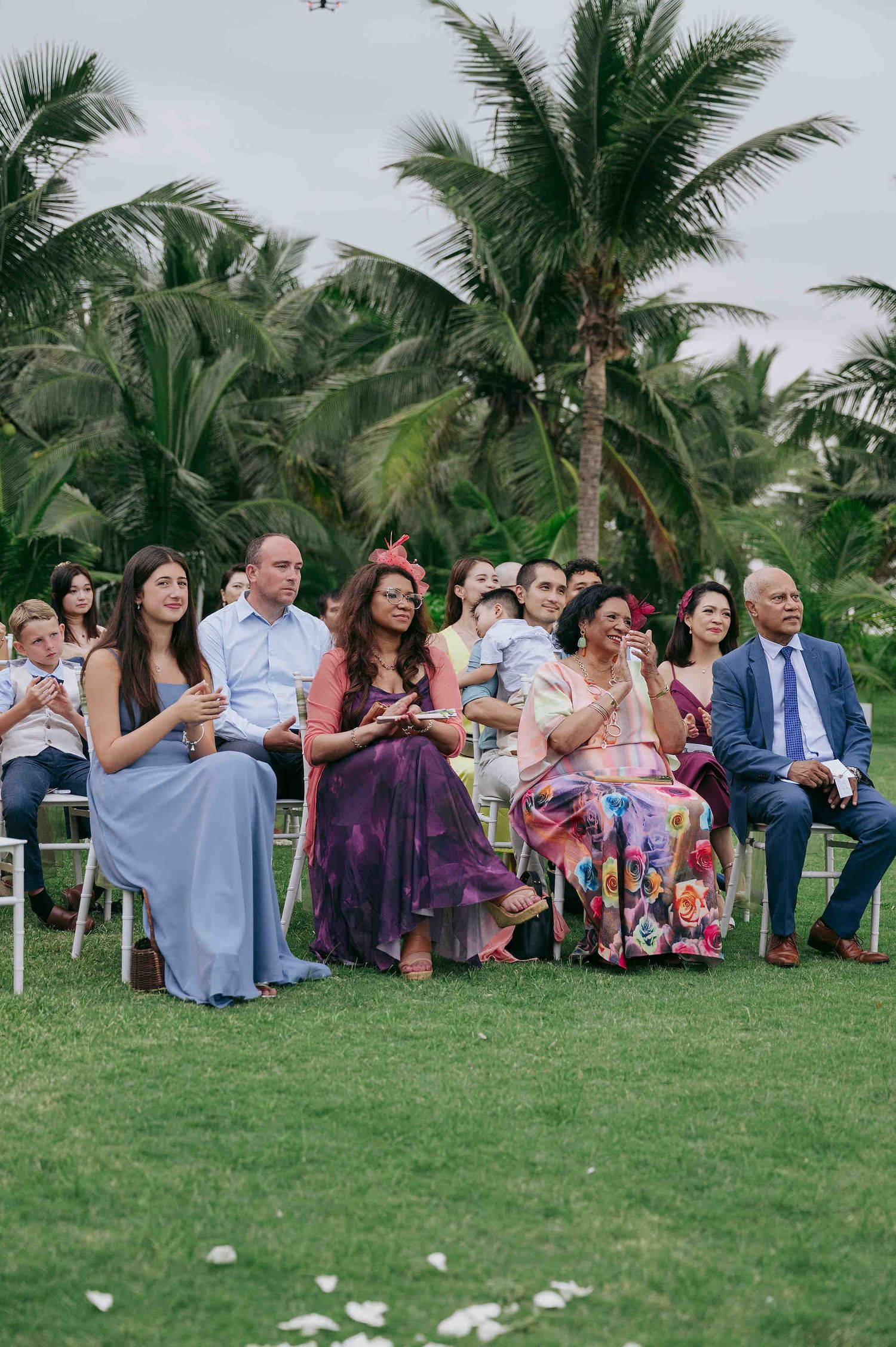 Wedding guests seated outdoors on green lawn, wearing colorful formal attire, with tropical palm trees in the background.