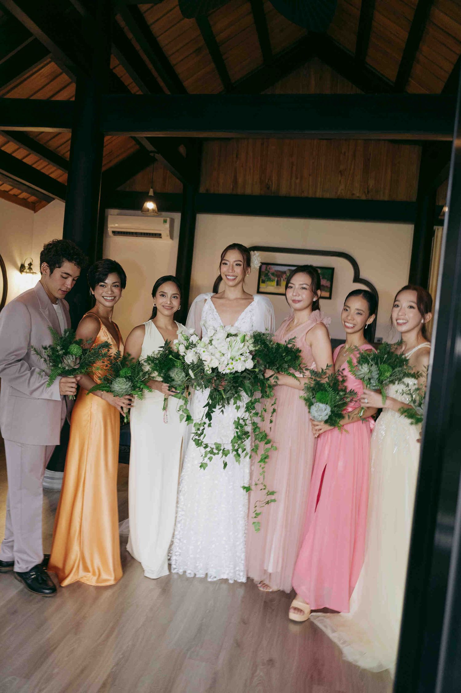 Bride in white lace gown poses with bridesmaids in colorful dresses holding green bouquets indoors.