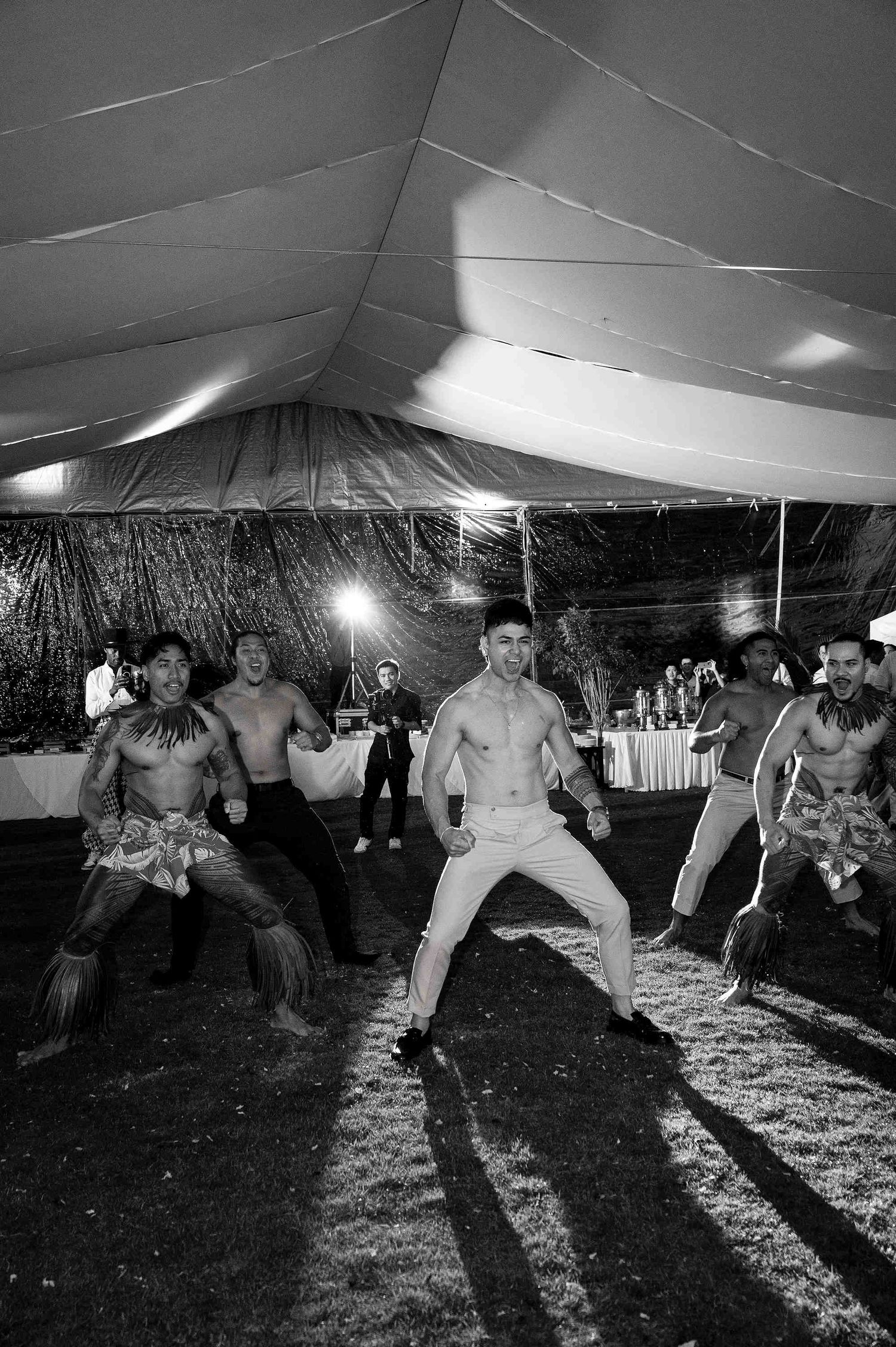 Black and white photo of shirtless male dancers performing a traditional Polynesian dance under a tent at an outdoor event.
