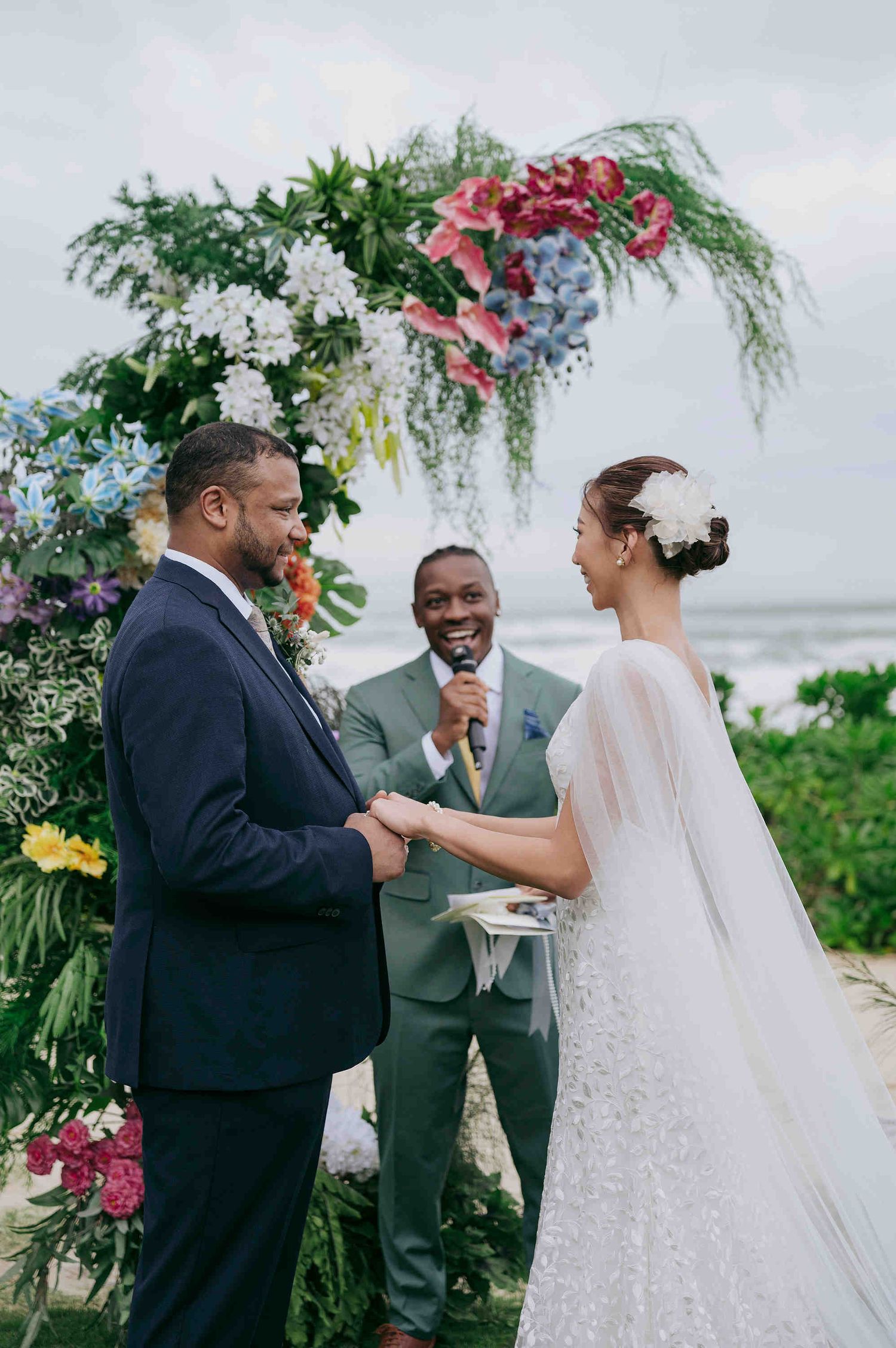 Couple exchanging vows at a tropical beach wedding ceremony with colorful floral arch and smiling officiant.