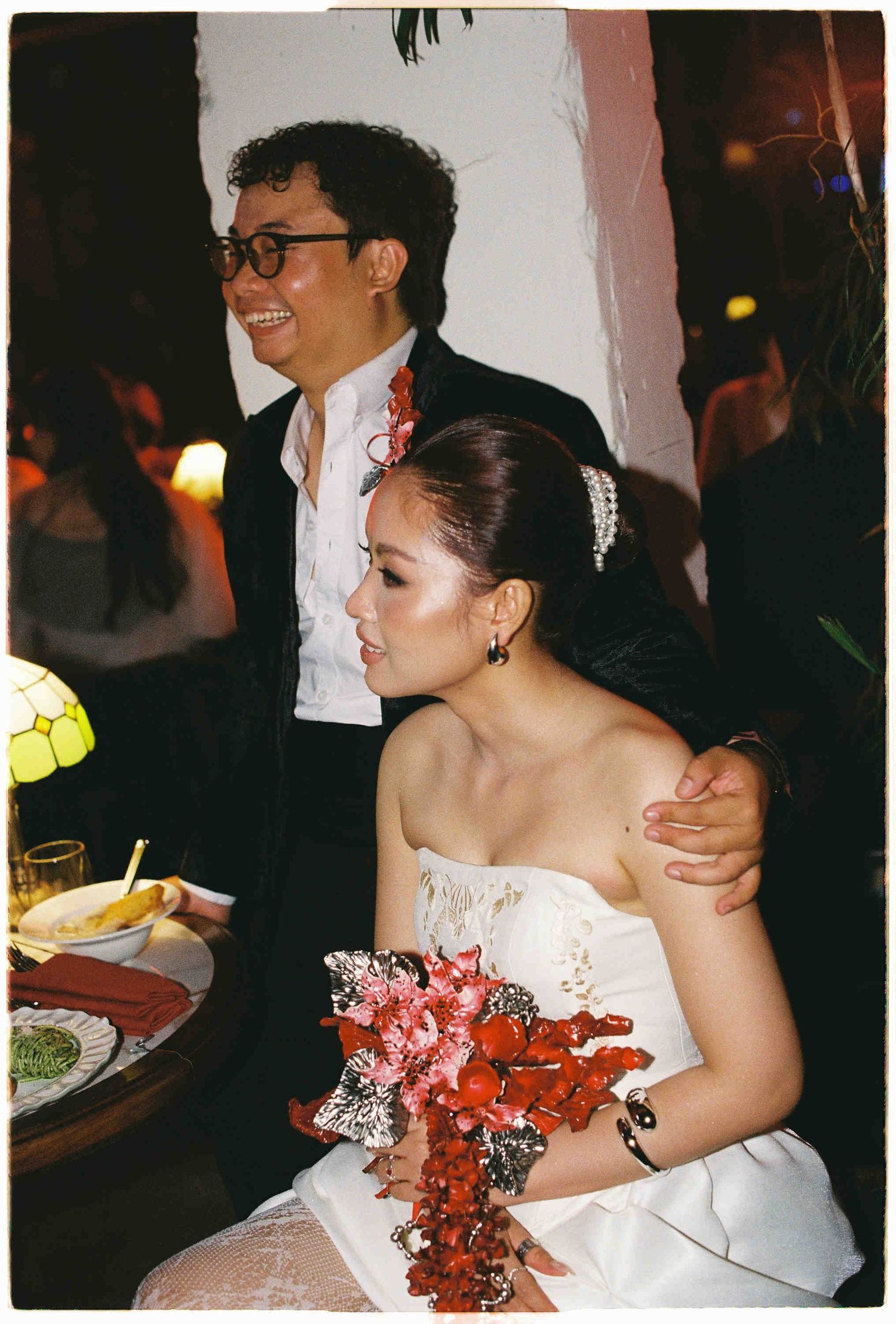 Bride in white strapless gown holding red floral bouquet sits beside groom in black suit at wedding reception.