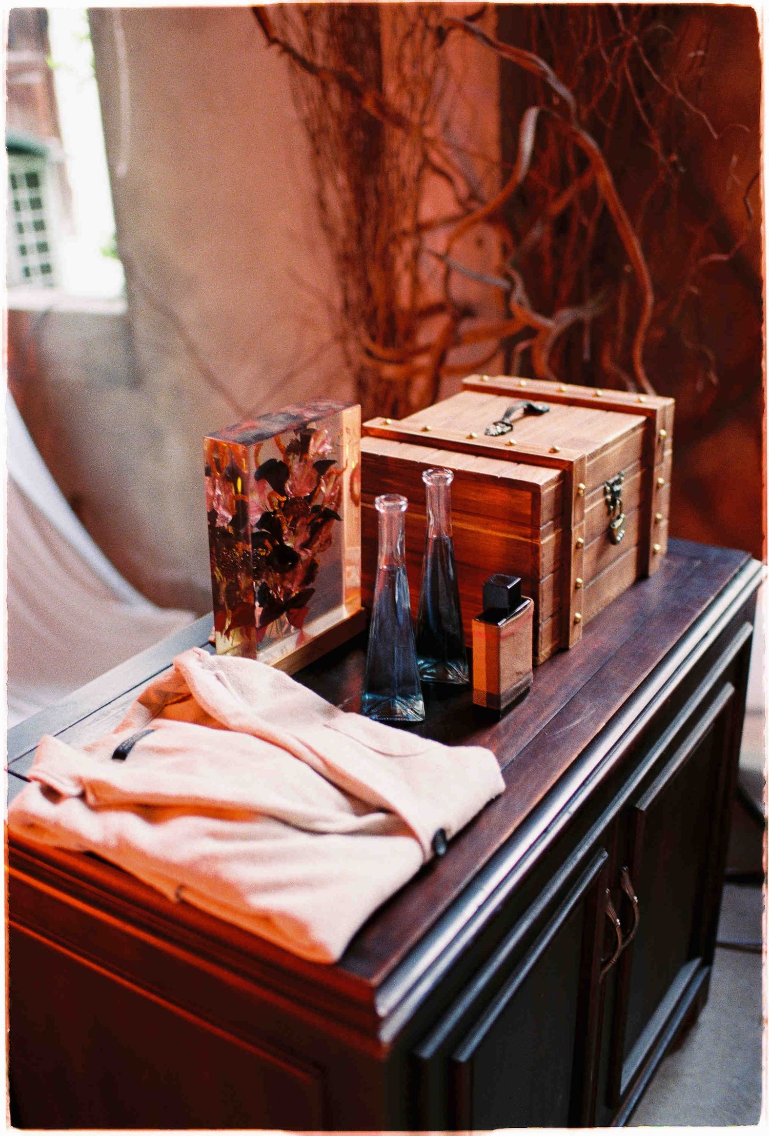 Rustic wooden chest box with glass bottles and pink fabric on dark wood dresser, decorated with dried branches.