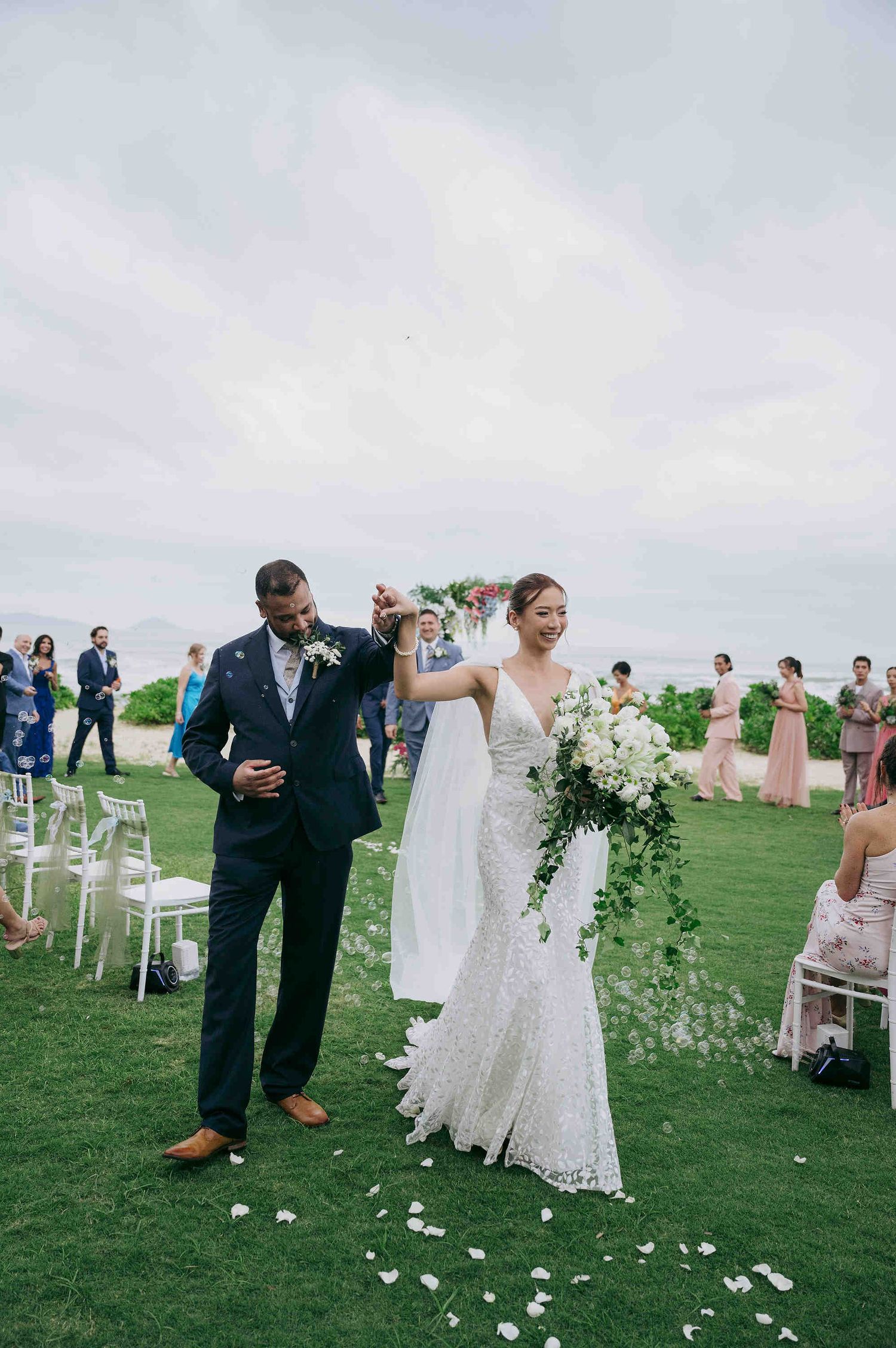 Bride and groom walk down outdoor aisle after wedding ceremony, bride holds white floral bouquet with greenery.