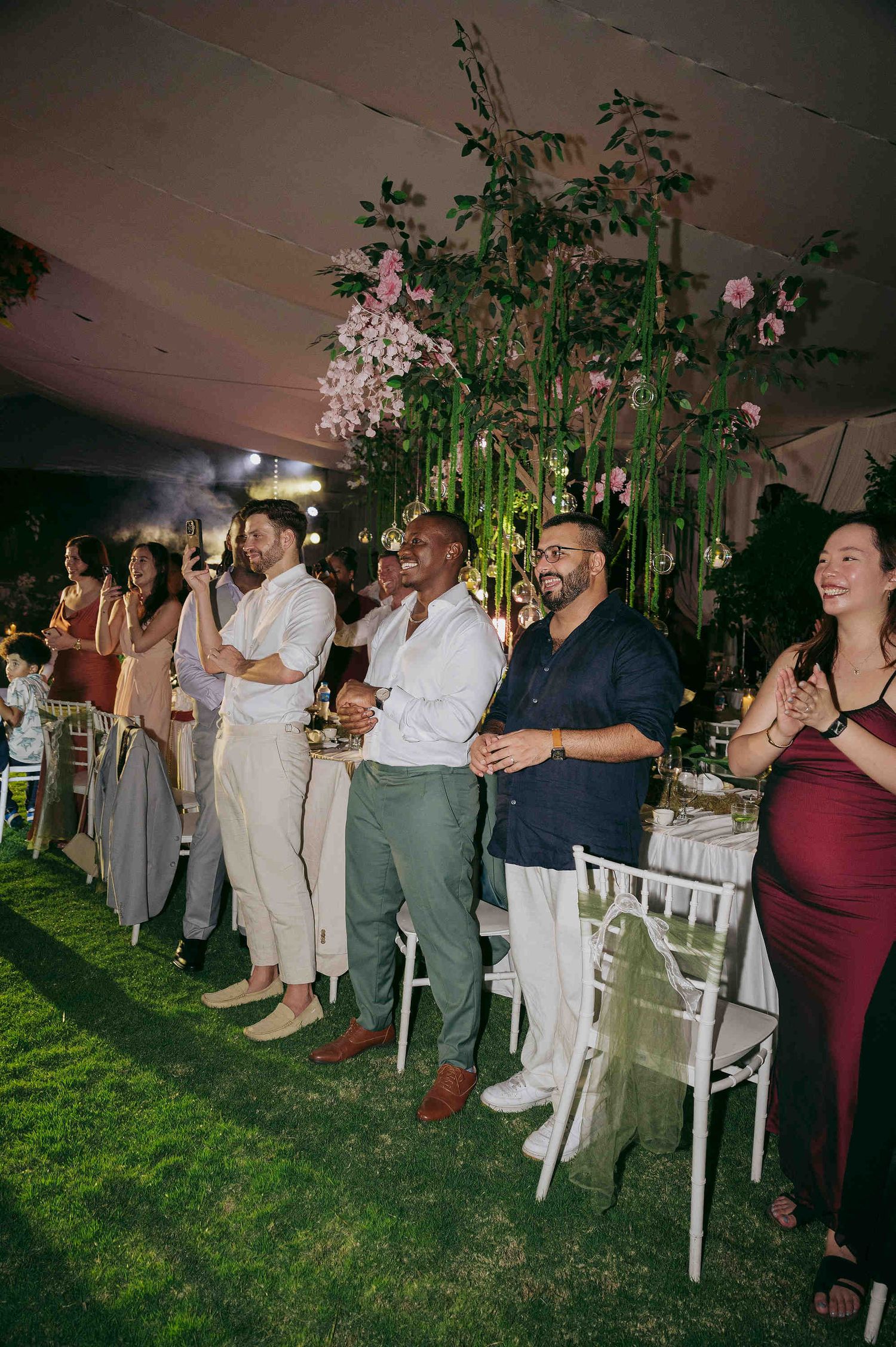 Guests stand and applaud at an elegant outdoor wedding reception under a tent with lush floral arrangements.