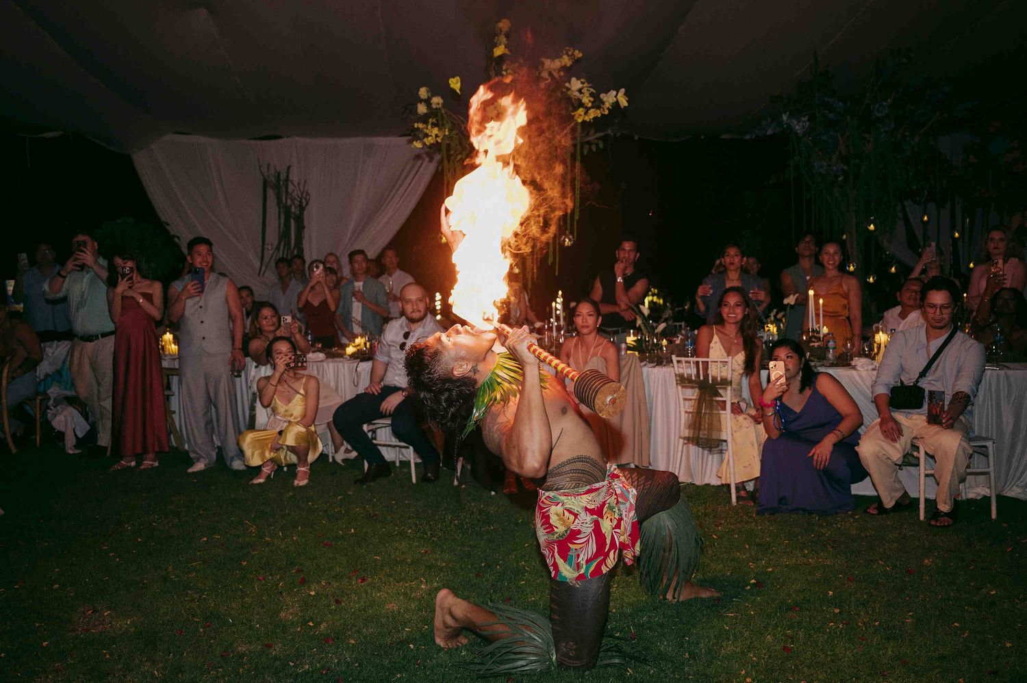 Fire breather performing at an outdoor evening event with crowd watching under a tent.