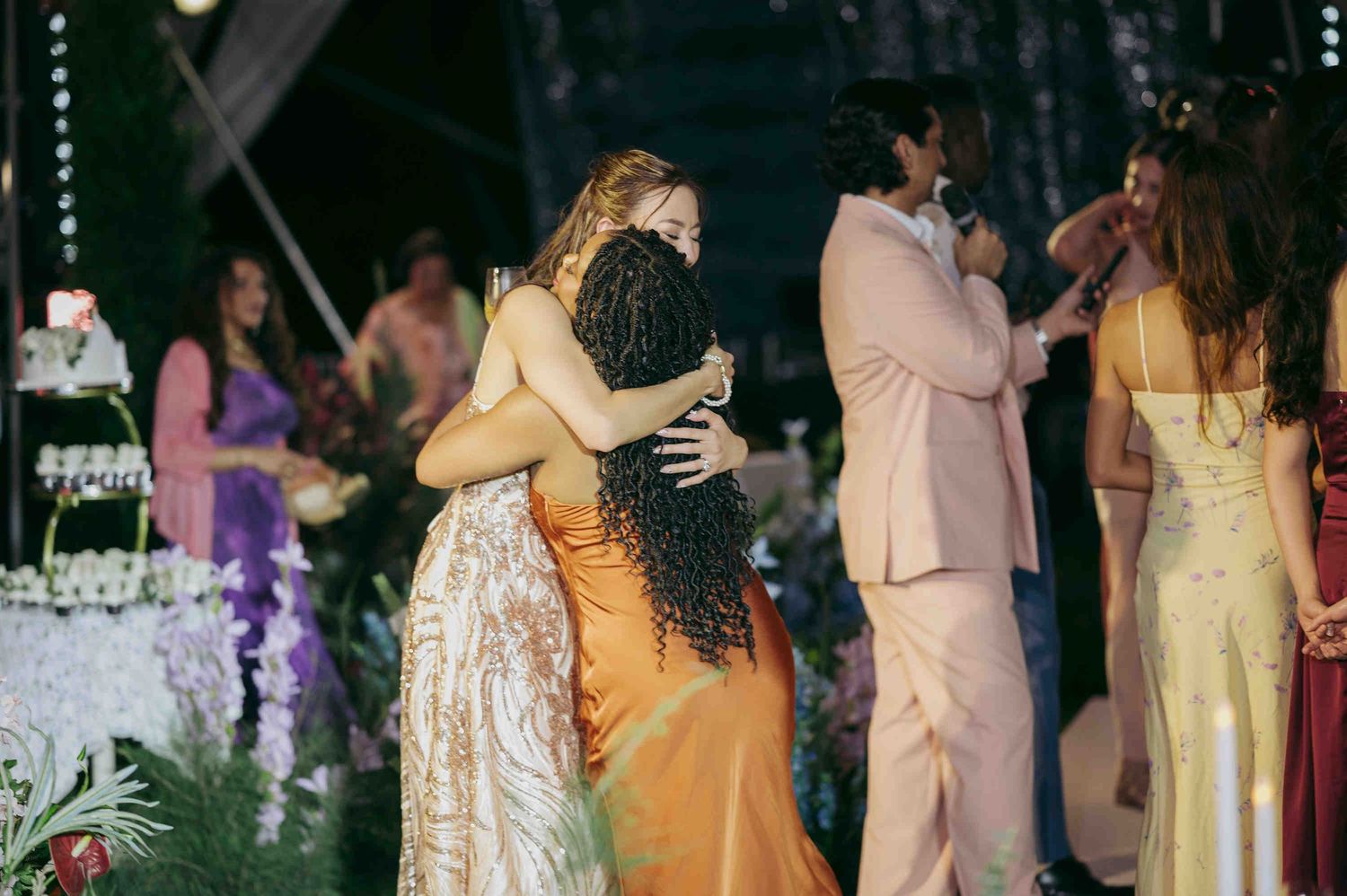 Two women embrace warmly at an elegant outdoor evening event, surrounded by guests in formal attire.