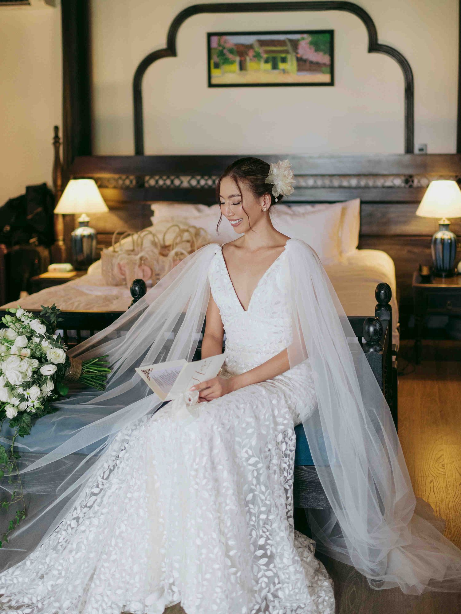 Bride in lace wedding gown with veil sits gracefully in elegant hotel room holding white floral bouquet.