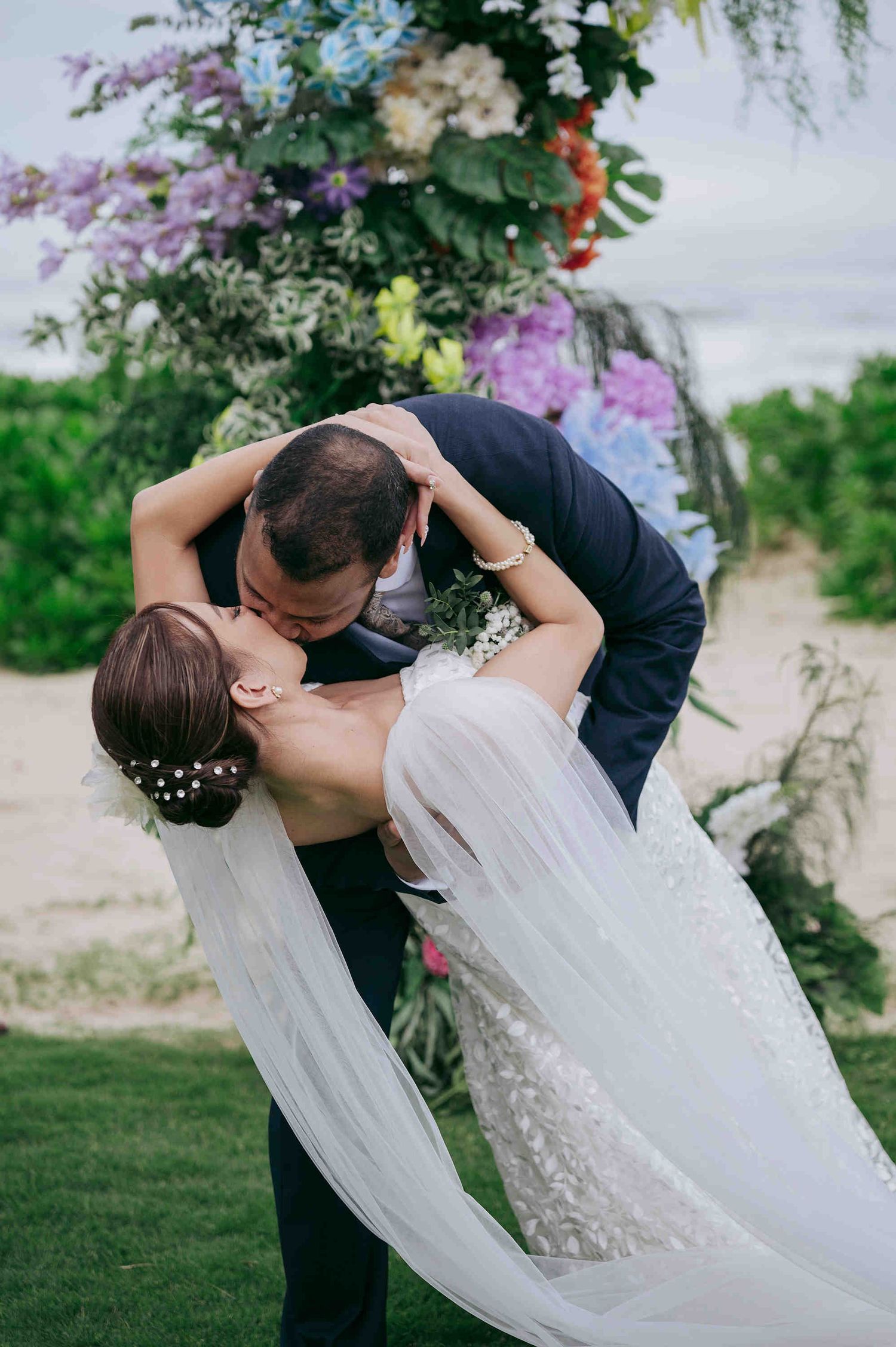 Bride and groom share a romantic dip kiss at outdoor beach wedding ceremony with colorful floral arch backdrop.