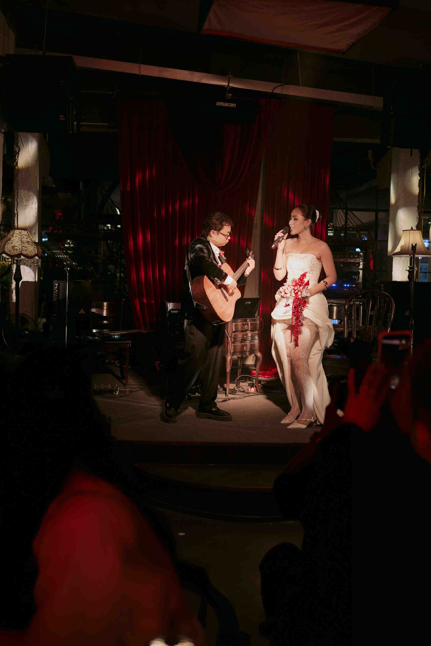 A guitarist and female singer in white dress perform on a dimly lit stage with red curtains backdrop.