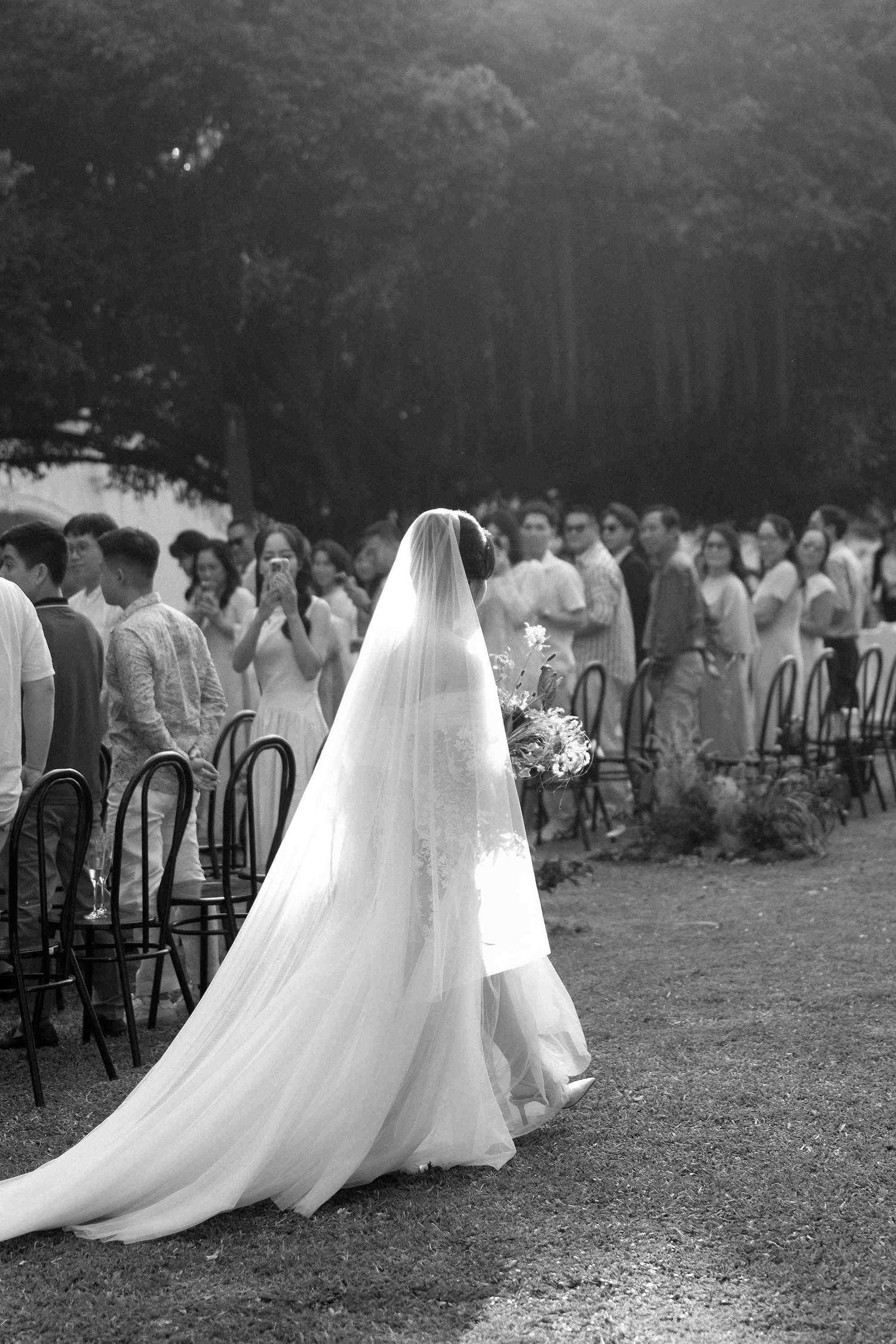 Black and white photo of a bride in a long veil walking down the aisle at an outdoor wedding ceremony.