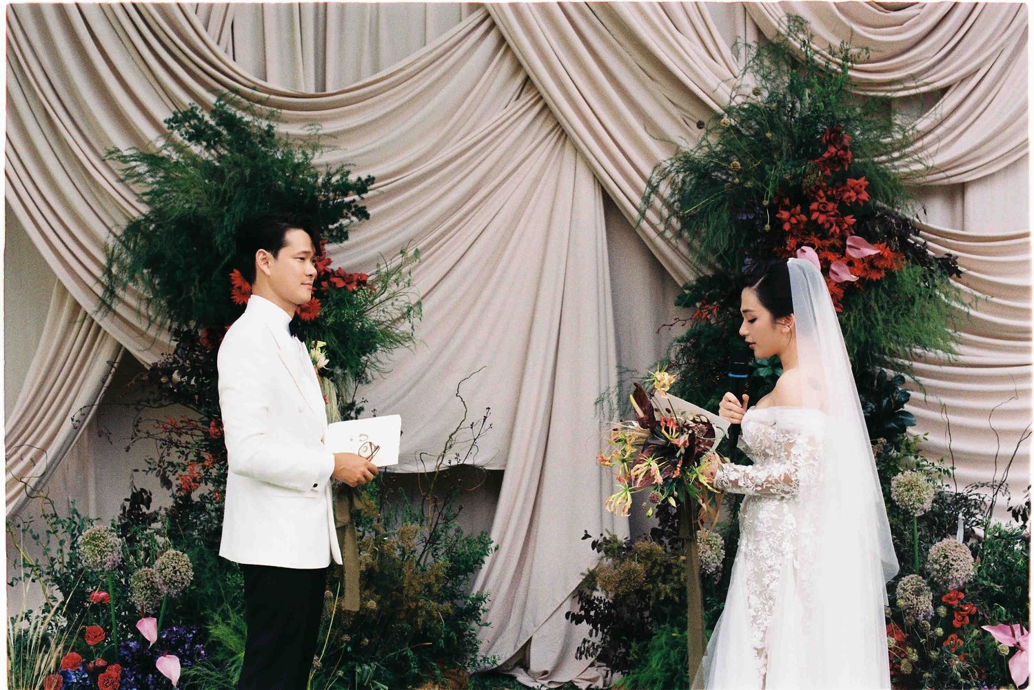 Bride and groom exchange vows at elegant wedding ceremony with lush floral backdrop and draped fabric decor.