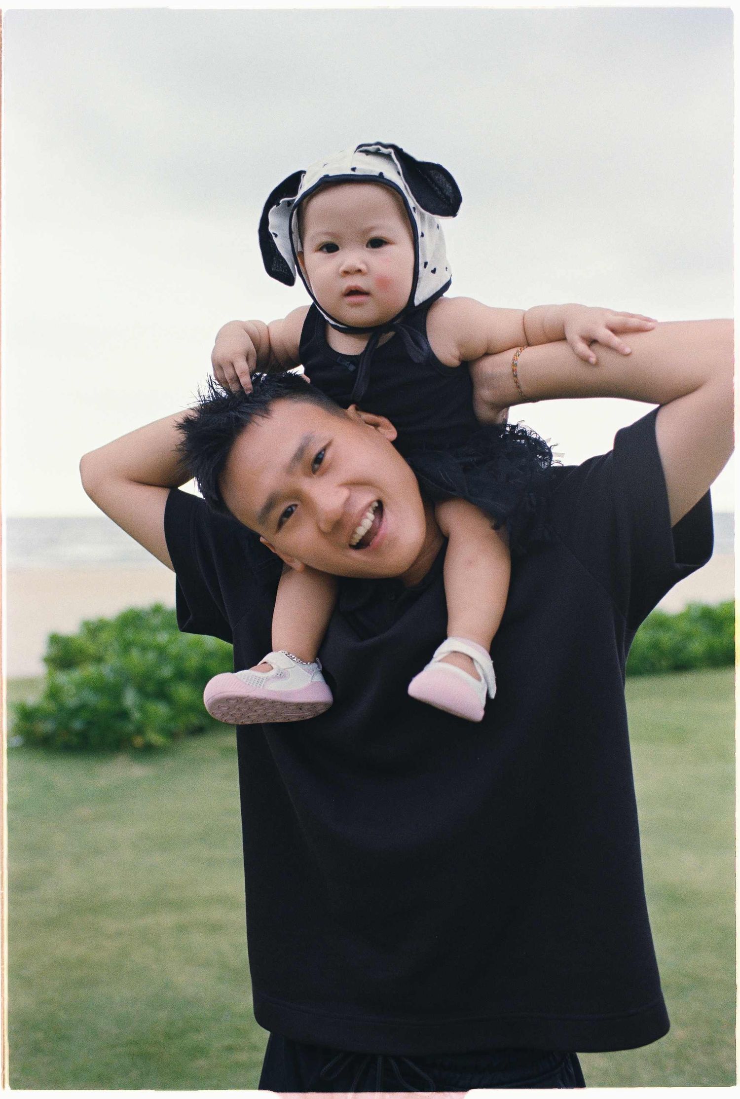 Da Nang family: playful moment captured outdoors as a baby wearing a hat sits on shoulders, by AnhVuWedding.