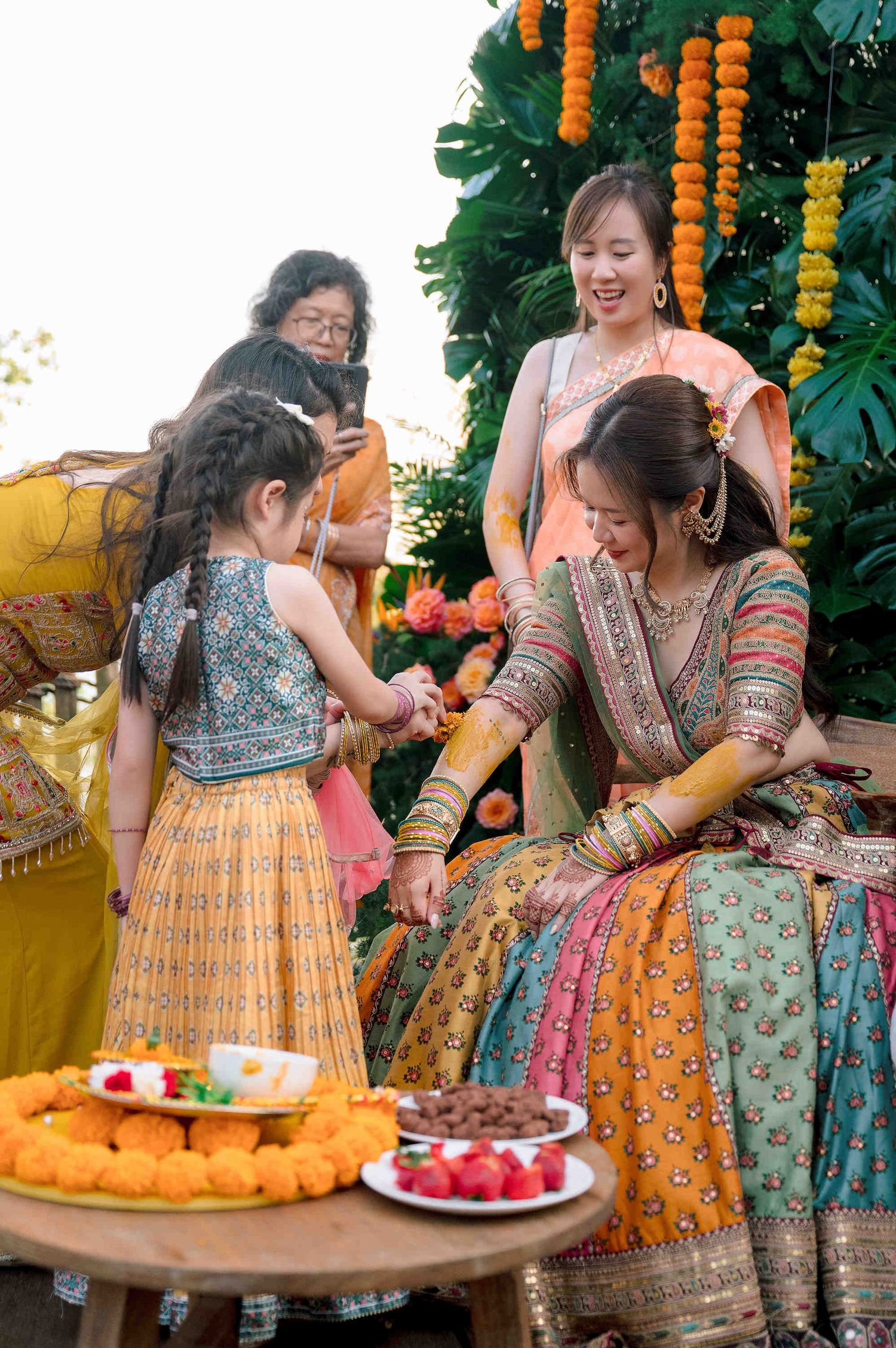 A bride in colorful Indian bridal attire receives henna during a traditional Mehndi ceremony surrounded by smiling guests.
