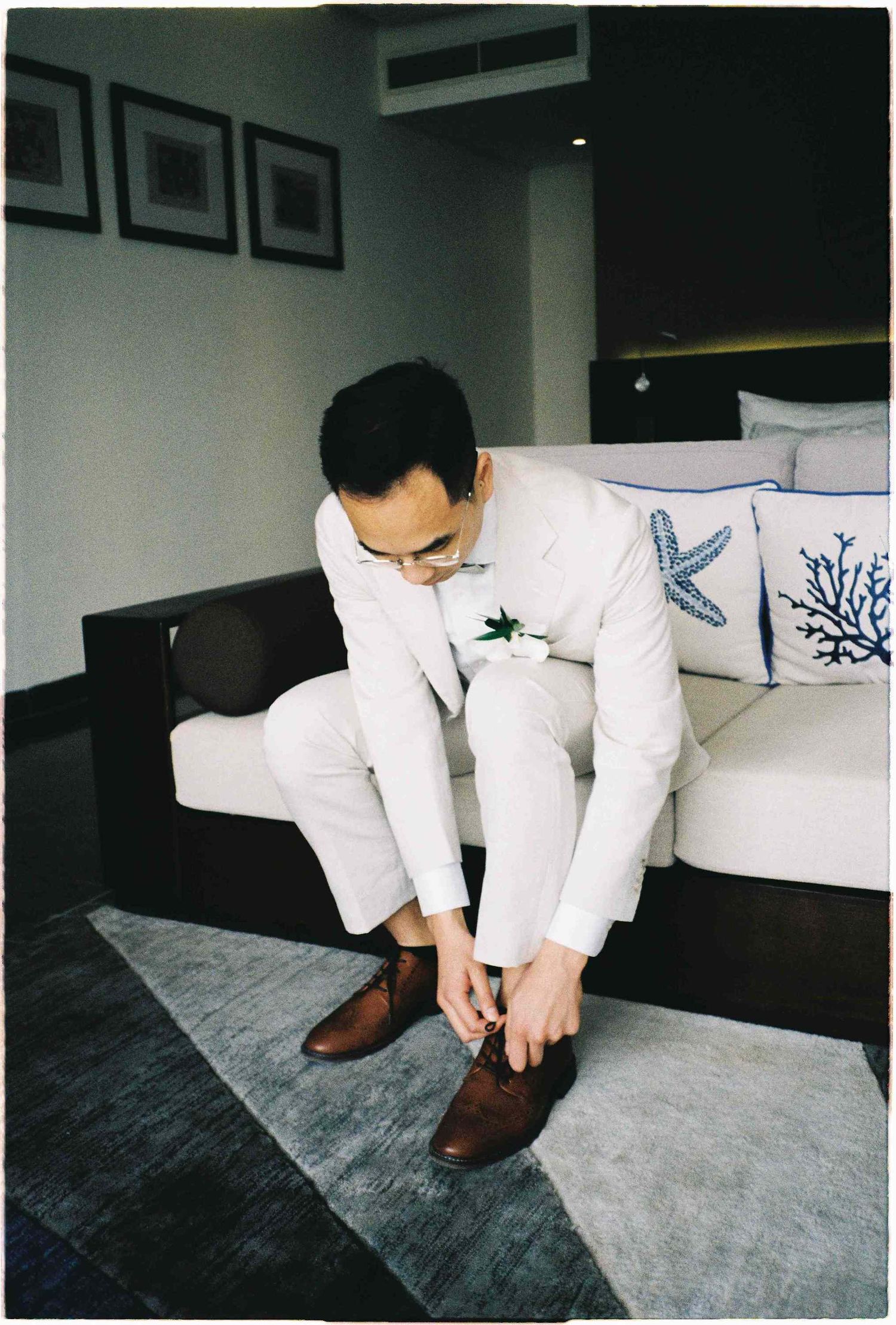 Groom in white suit with boutonniere tying brown dress shoes while seated on a white sofa in a hotel room.