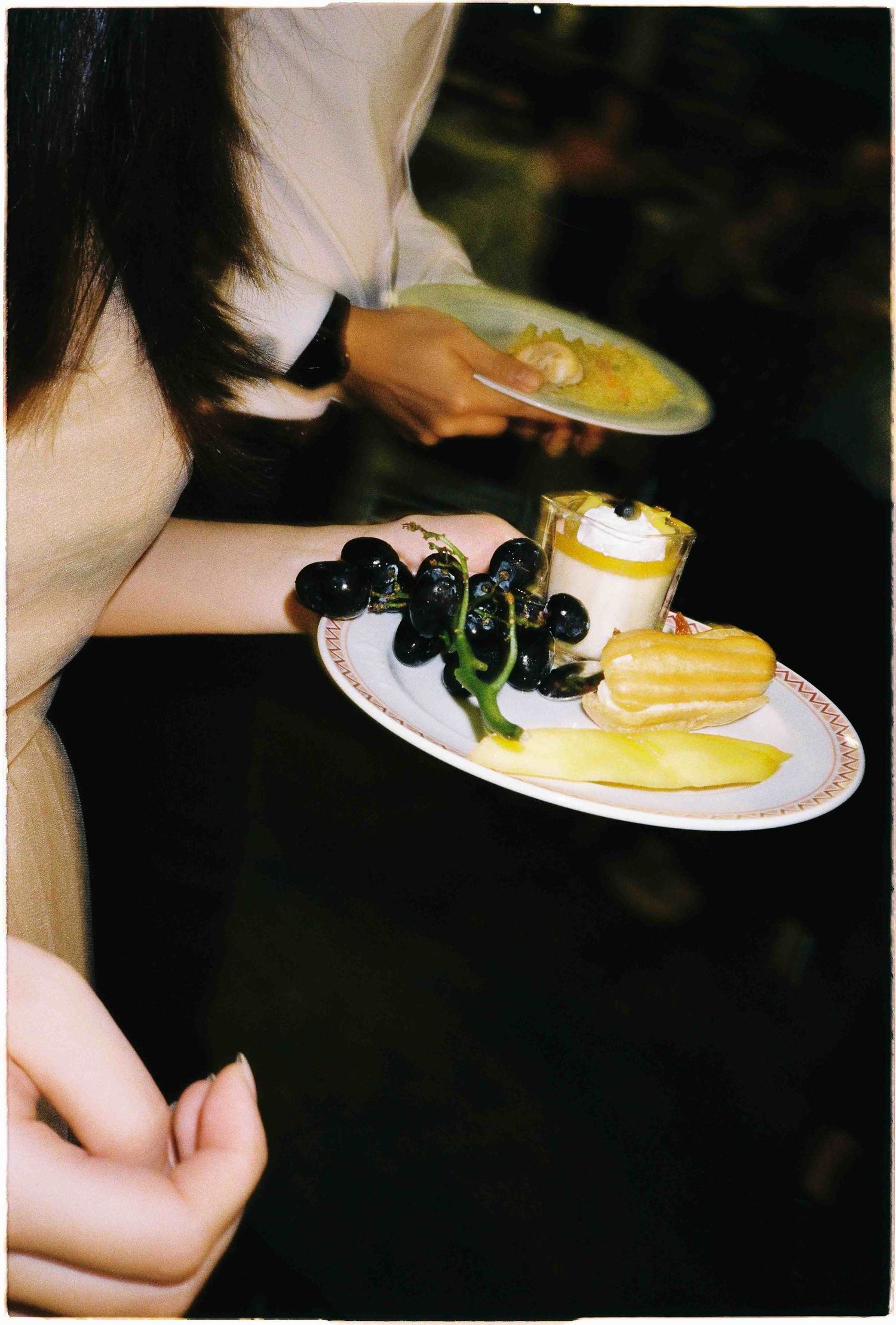 A woman holds a decorative plate with grapes, eclairs, and a cream dessert at a party.