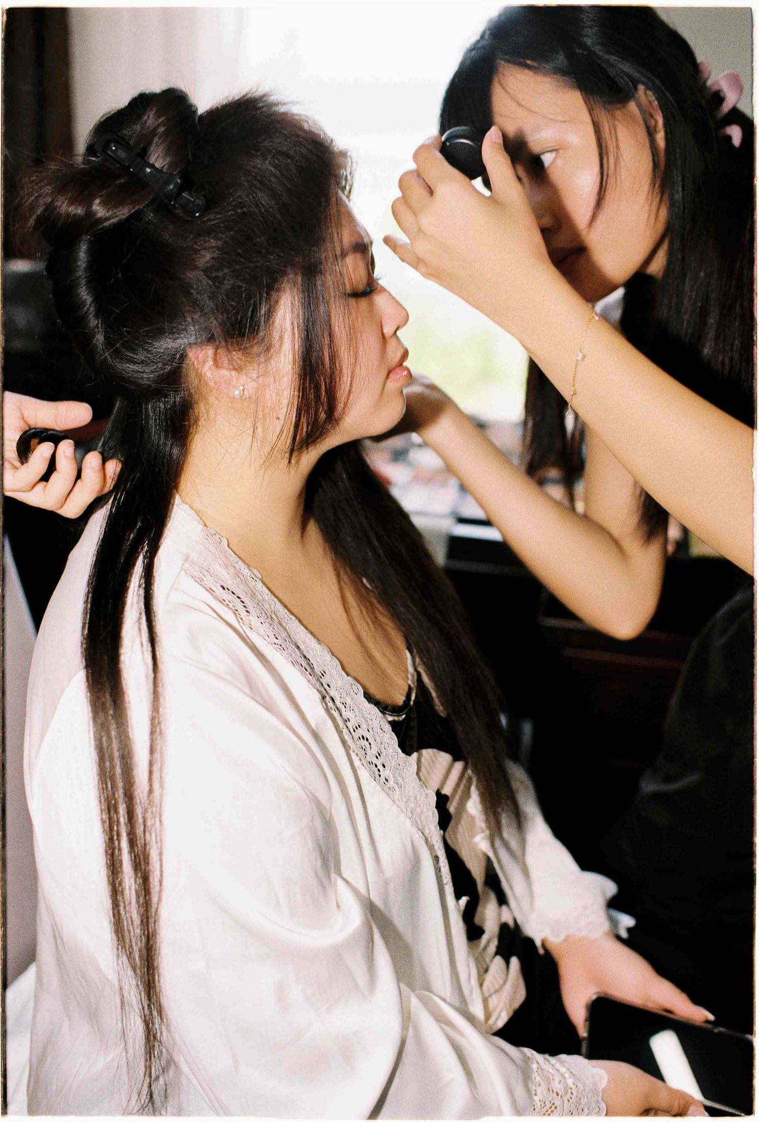 A makeup artist applying eye makeup to a bride in a white robe during wedding preparations.