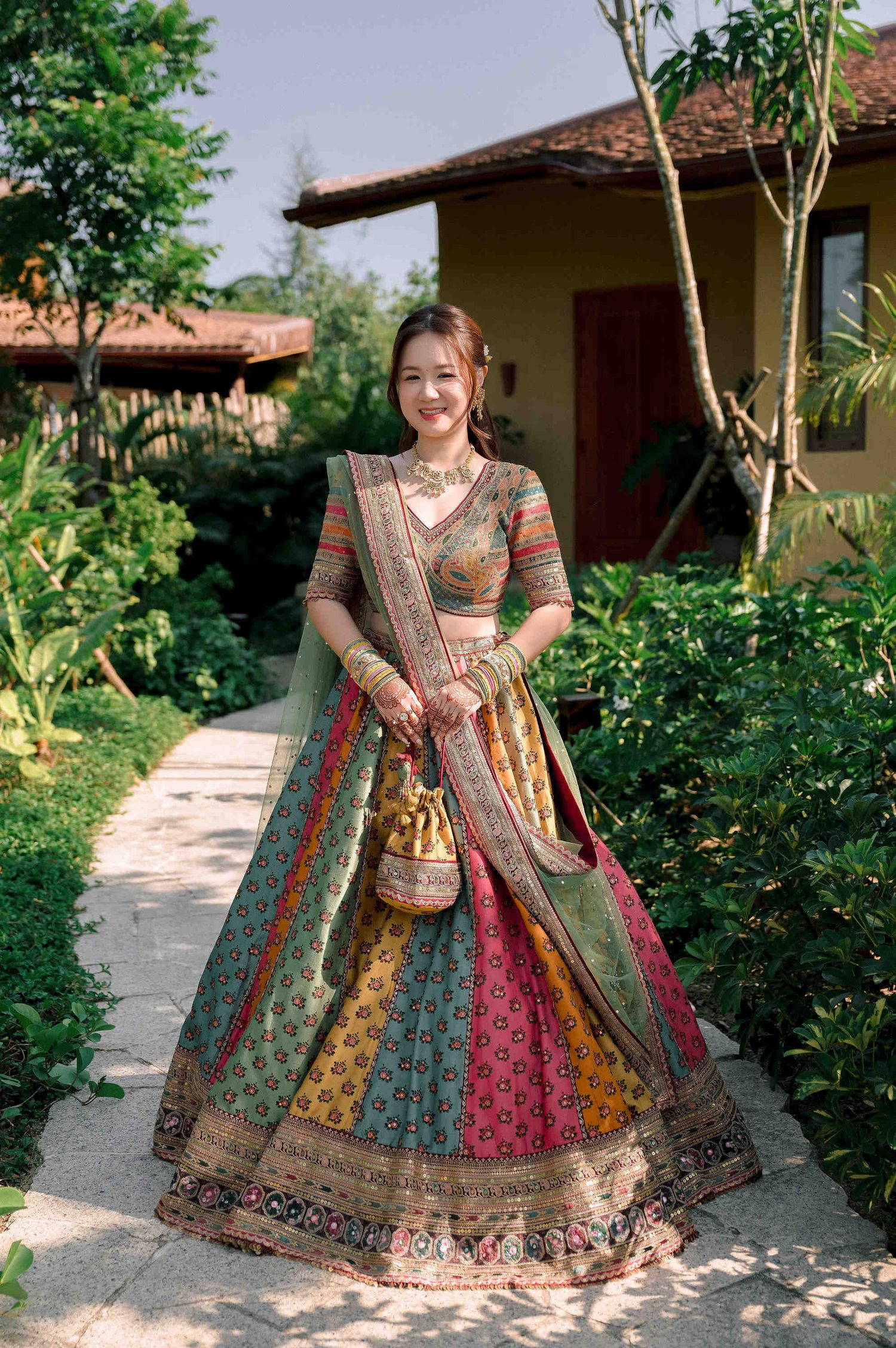 Woman smiling in colorful Indian lehenga choli with dupatta, standing on garden pathway near traditional buildings.