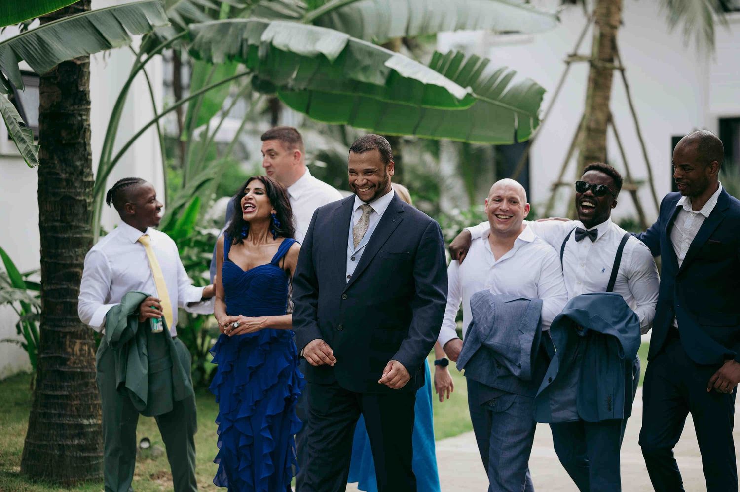 Group of elegantly dressed wedding guests walking outdoors near tropical palm trees and white building.