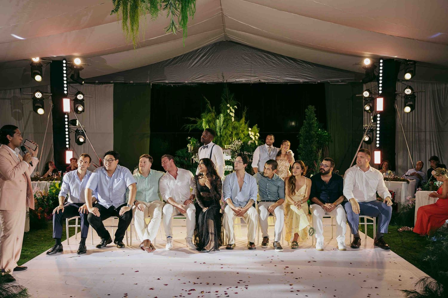Group of elegantly dressed guests seated in a row at an outdoor wedding reception under a decorated tent at night.