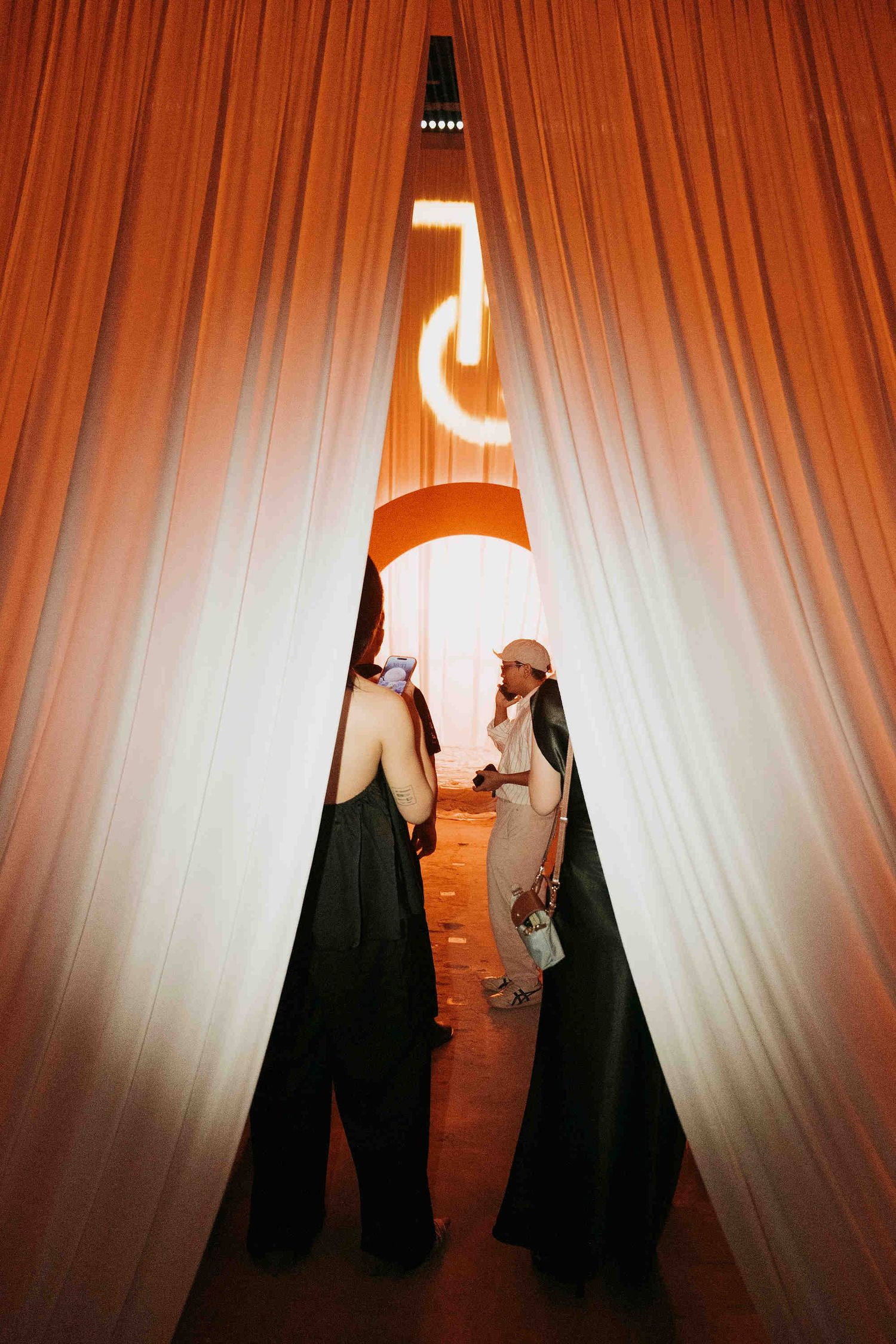 Two elegantly dressed women converse through parted orange curtains beneath a glowing neon sign at an upscale event.