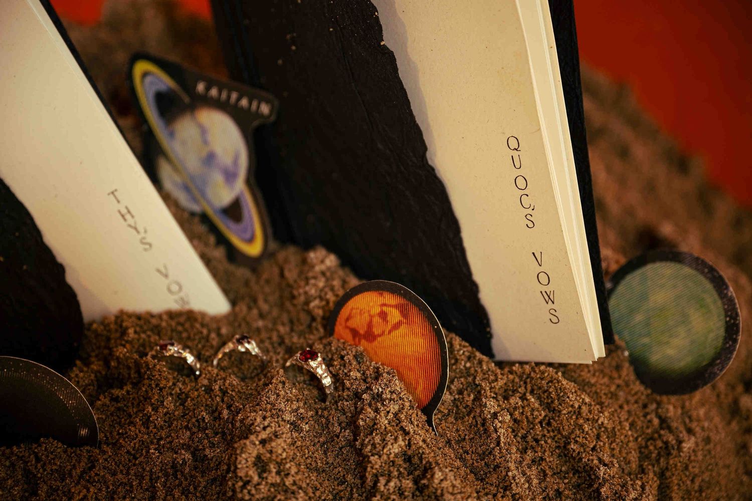 Books with planetary covers partially buried in sand, featuring Saturn and Mars imagery in a cosmic display.